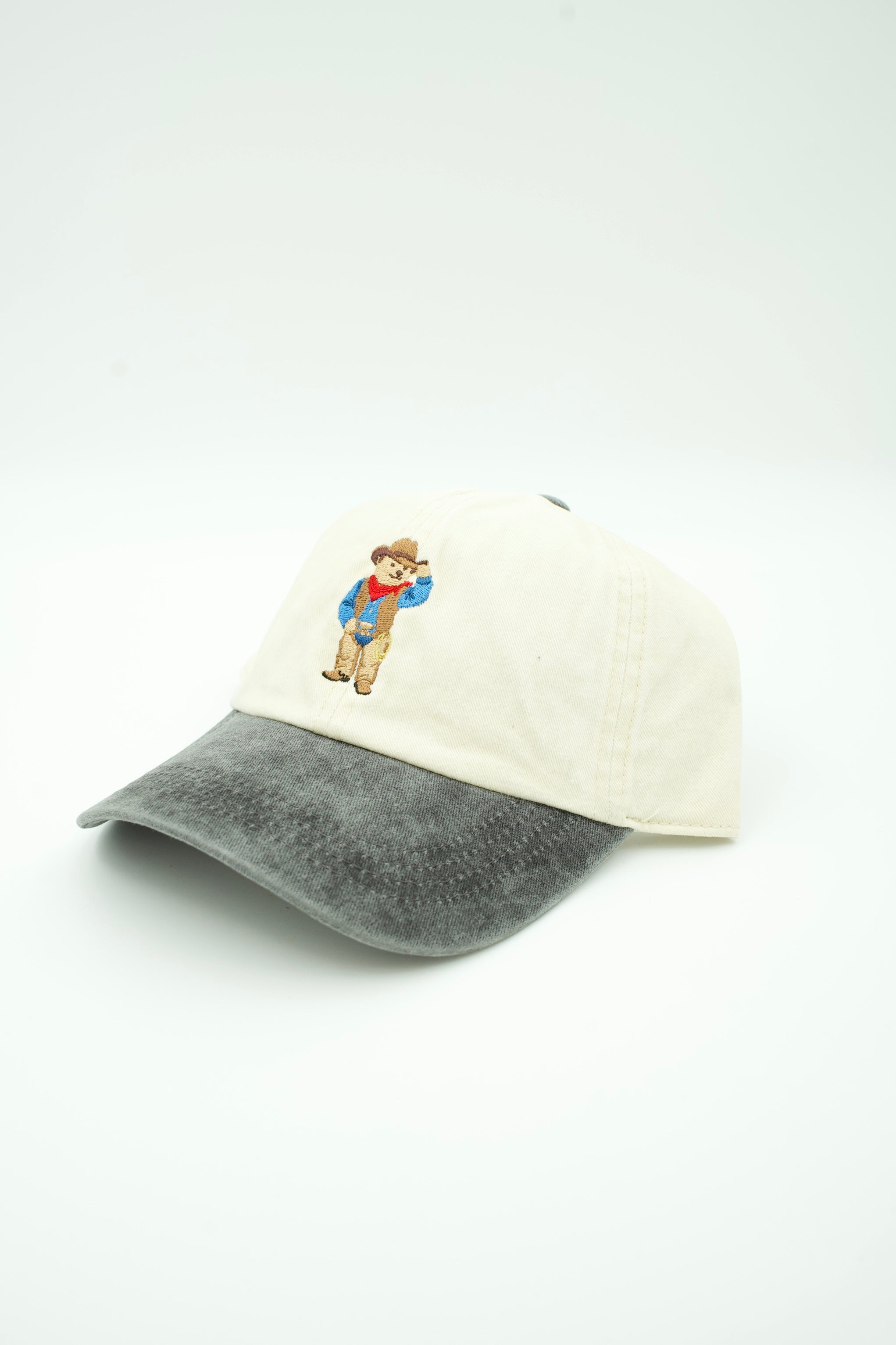 A baseball cap with an off-white crown and a dark gray washed brim, featuring a small embroidered cartoon boy on the front.