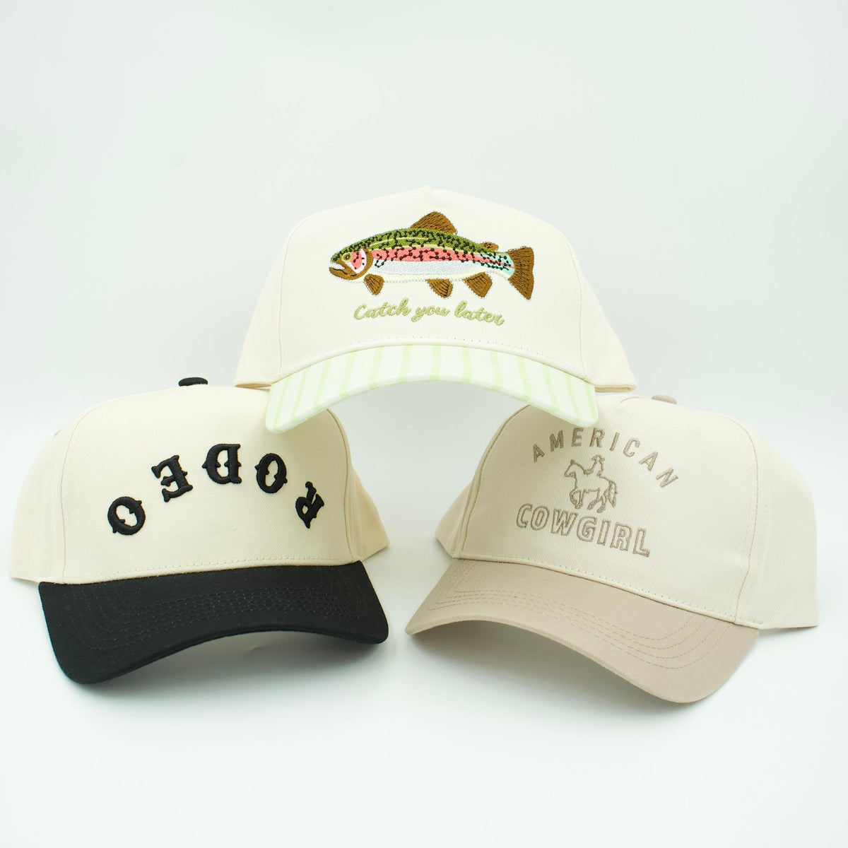 Three embroidered baseball caps displayed against a plain light background: one cream cap with a black brim and black 'RODEO' lettering, one beige cap with a black brim and tan 'American Cowboy' text, and one cream cap with a green-and-white striped brim and a multicolor fish patch reading 'Catch Your Fines.'.
