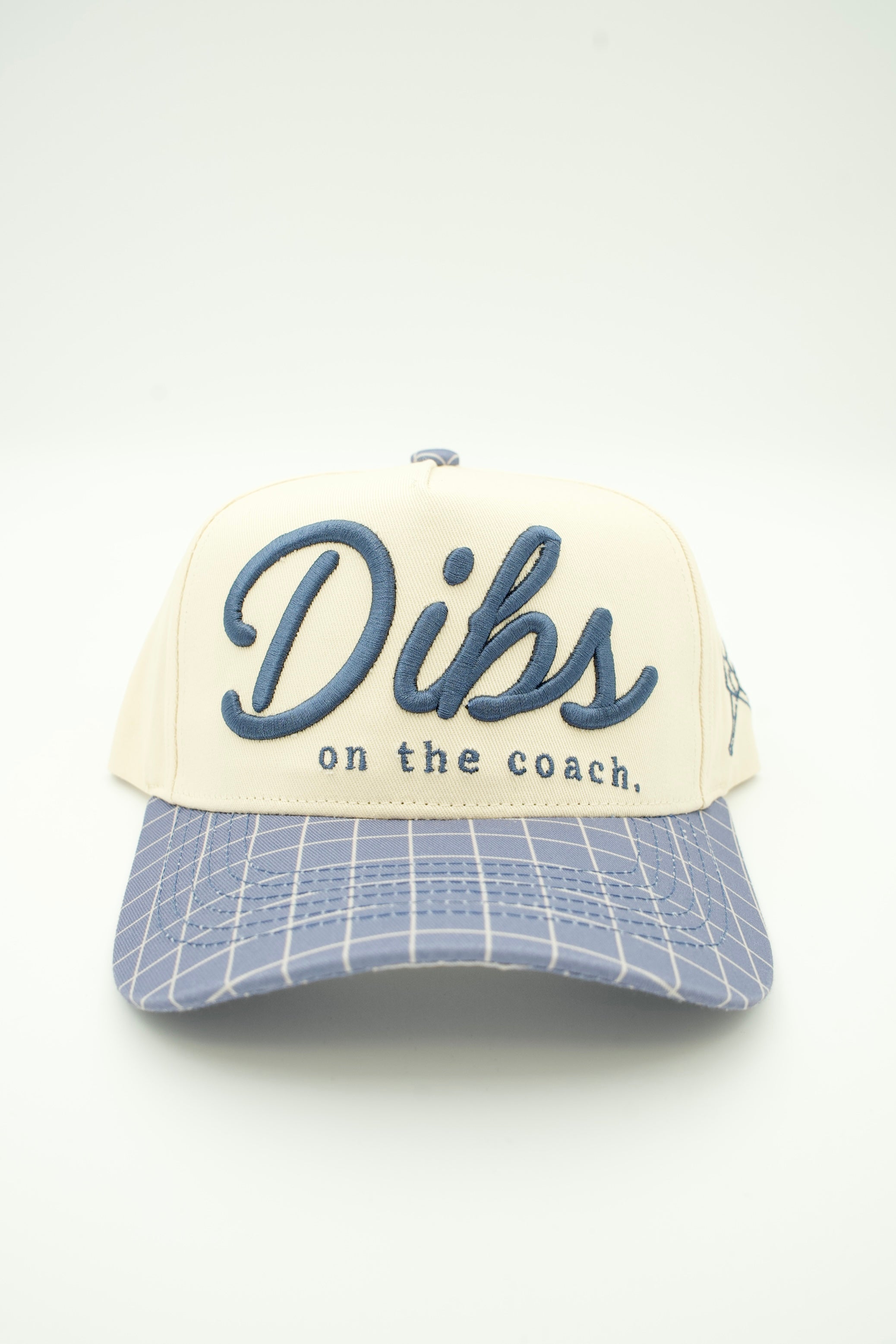 A trucker hat with an off-white crown, blue embroidered script reading 'Dibs on the coach.', and a light blue and white plaid brim, shown against a plain white background.