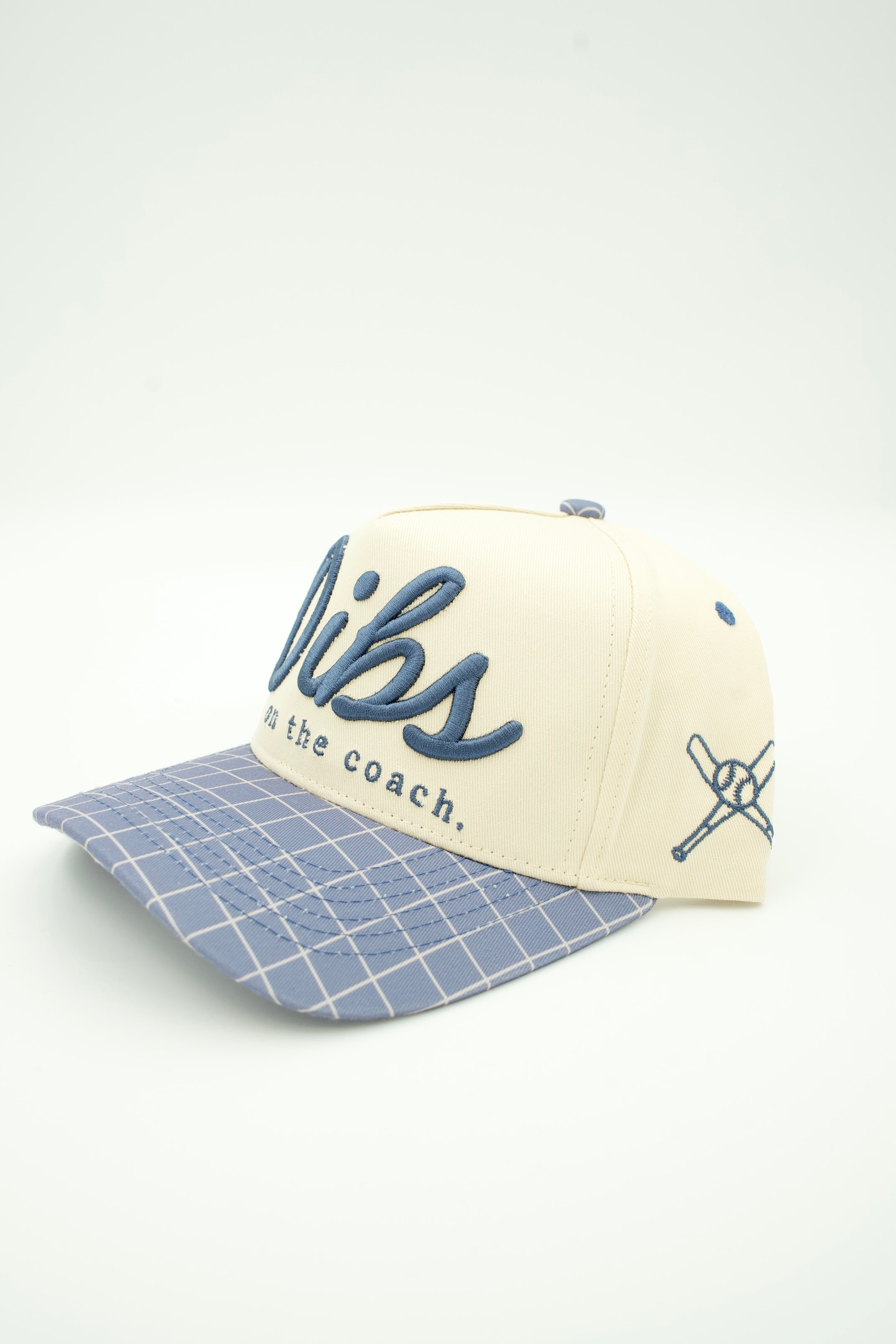 A baseball cap with a cream crown, navy blue script lettering on the front, and a light blue and white grid-patterned visor, shown on a white background.