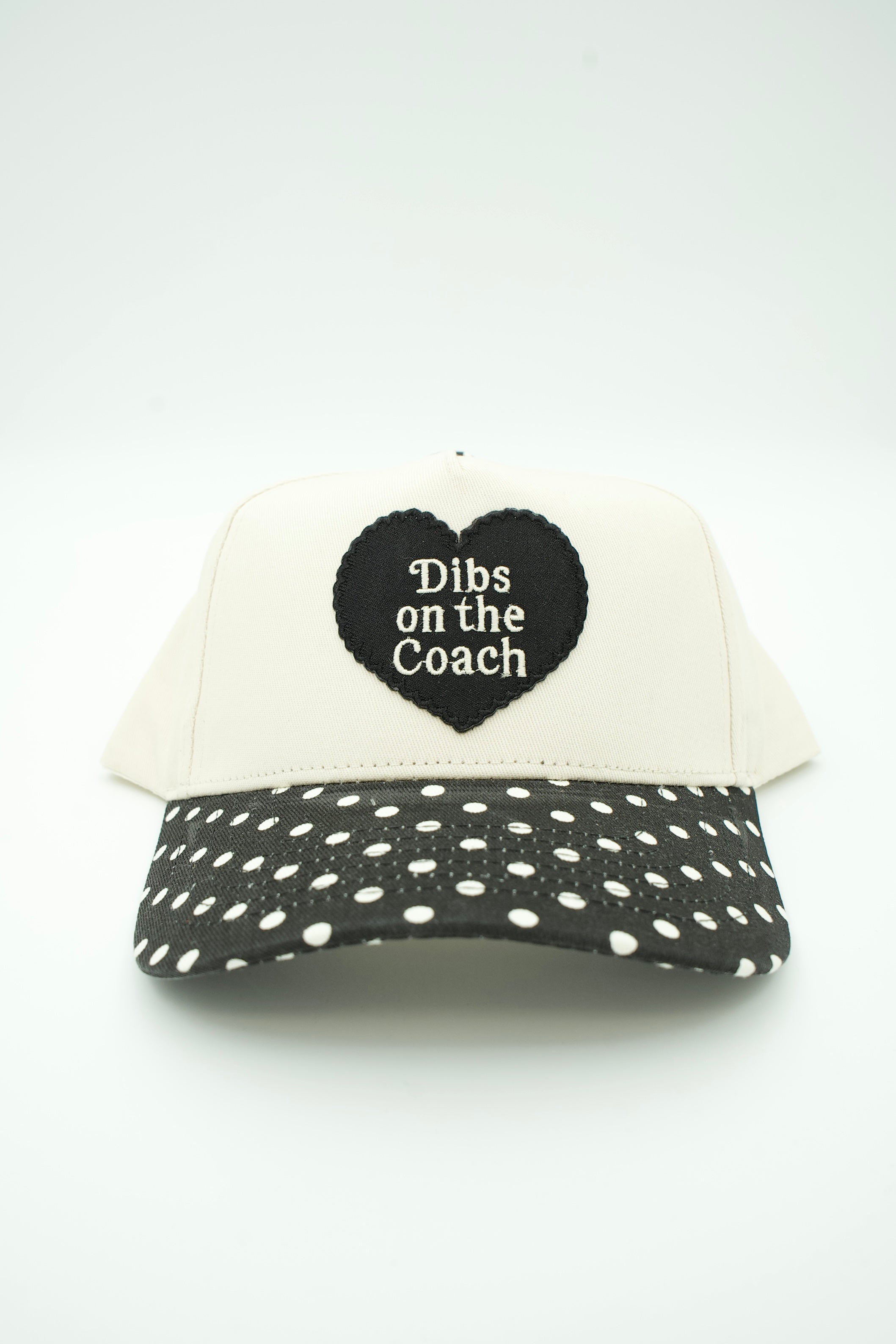 A baseball cap with an off-white crown and a black brim covered in white polka dots. The front features a black heart patch with the text 'Dibs on the Coach' in white.