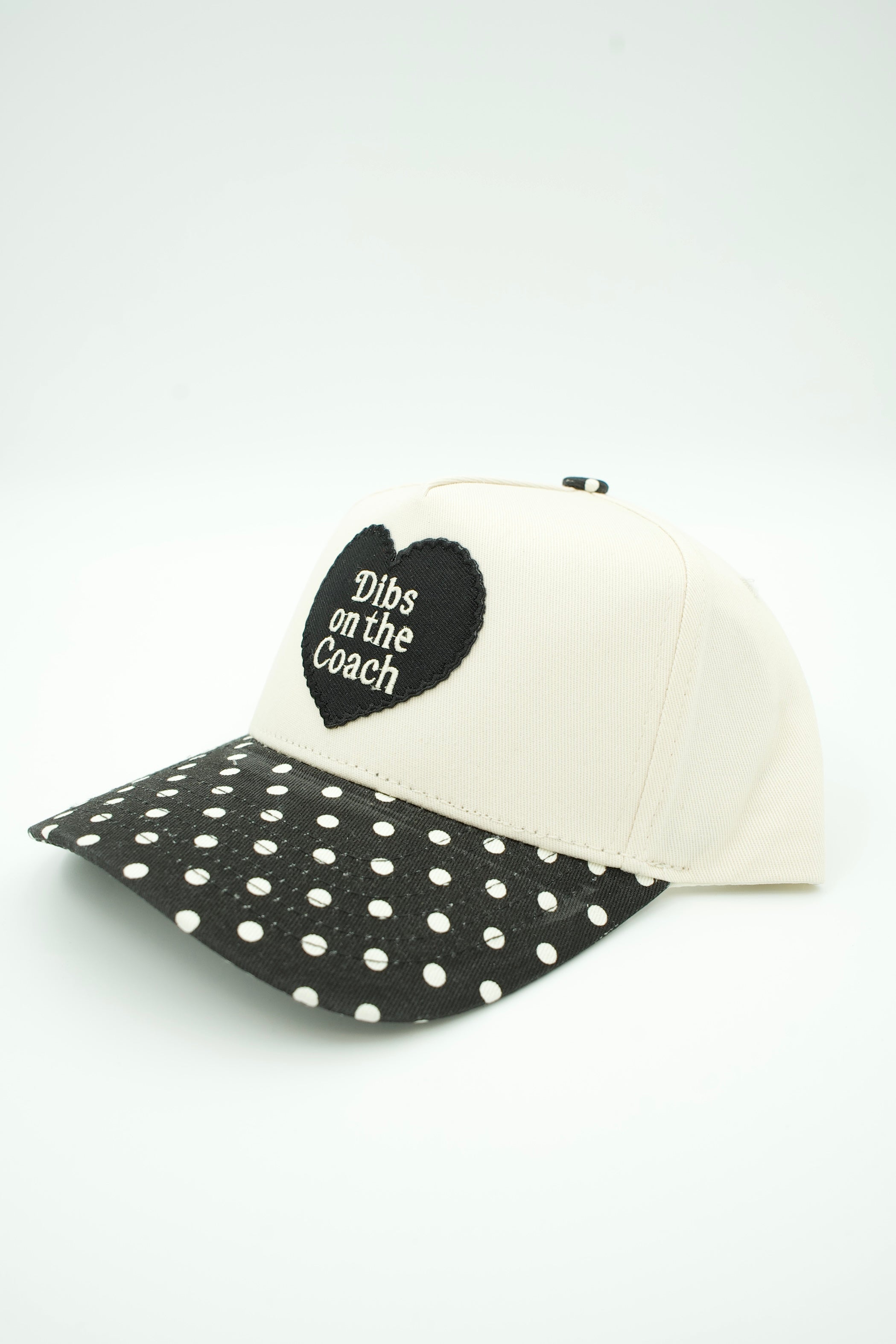 A baseball cap with an off-white crown and a black brim covered in white polka dots. A black heart patch on the front reads 'Dibs on the Coach'.