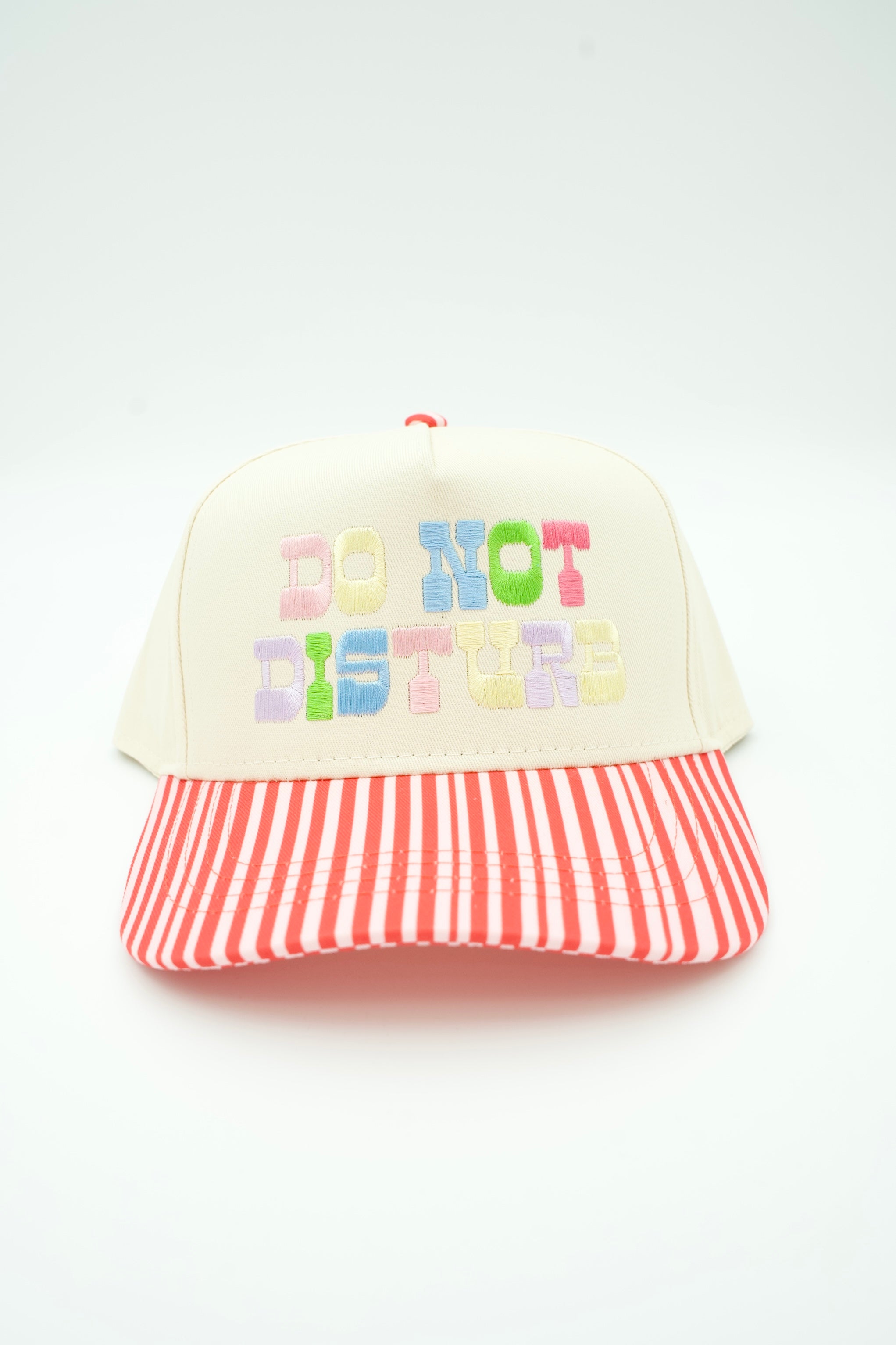 A cream-colored trucker-style cap with a red and white striped brim. The front panels feature the embroidered text 'DO NOT DISTURB' in pastel-colored letters.