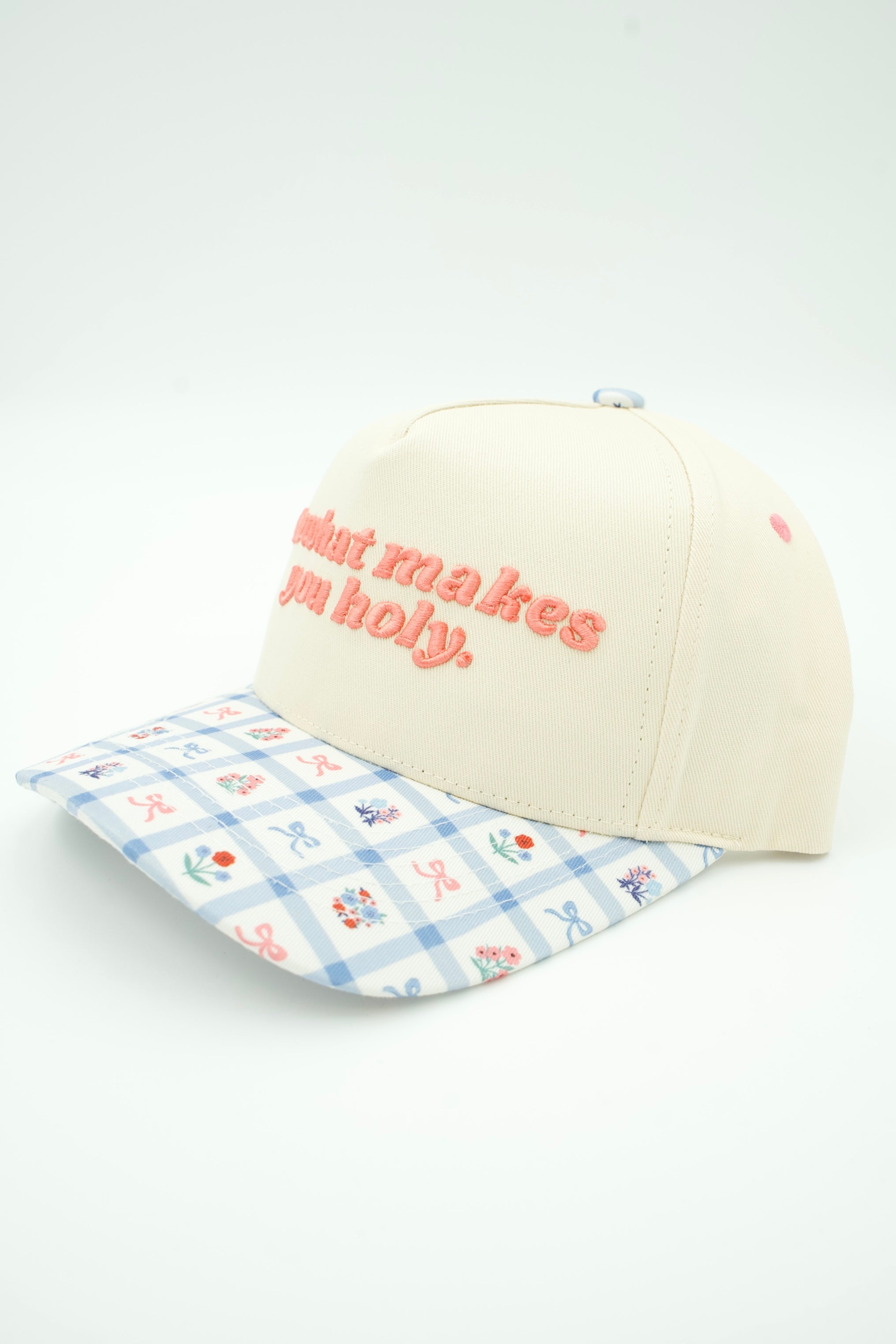 An off-white baseball cap with coral pink embroidered text reading 'do what makes you holy.' on the front, paired with a light blue and white gingham brim patterned with small pink and blue flowers.