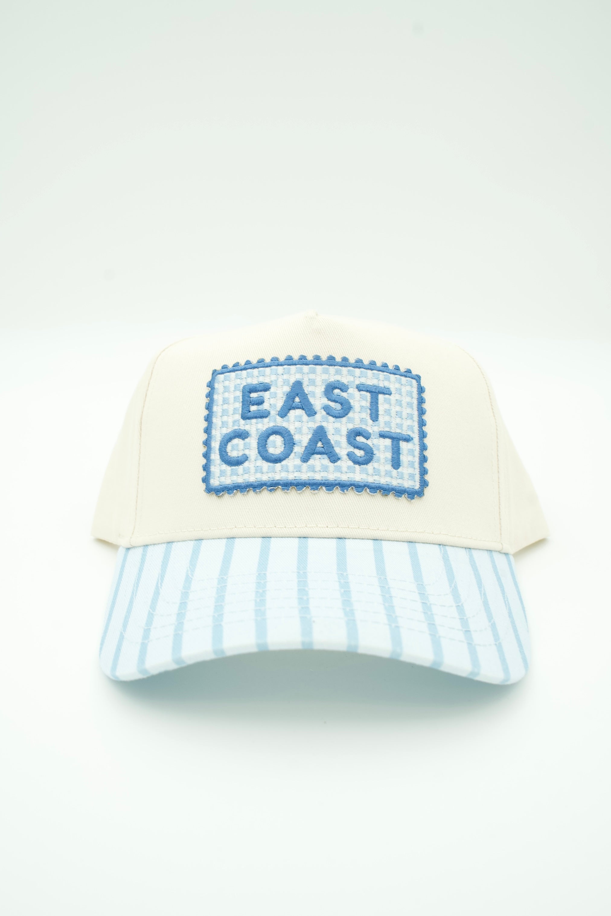 A baseball cap with an off-white crown, a light blue and white striped brim, and a blue embroidered patch on the front that reads 'EAST COAST'.