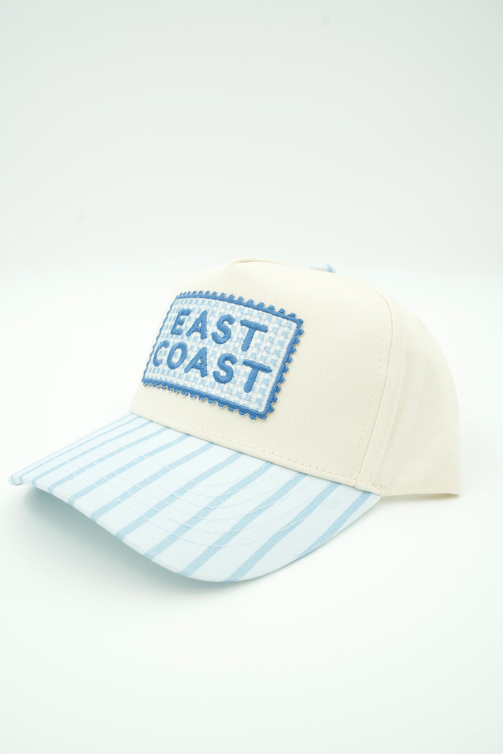 An off-white baseball cap with a light blue and white striped brim and a blue embroidered 'EAST COAST' patch on the front, set against a plain white background.