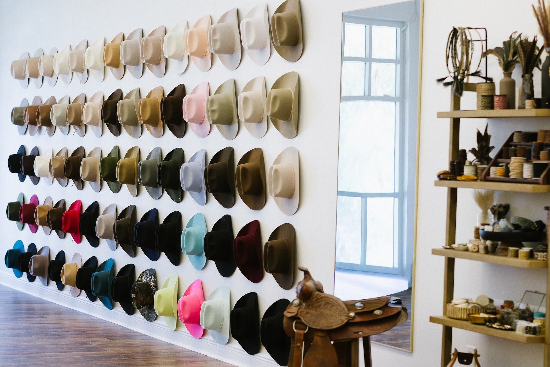 A large wall display of many cowboy hats in different colors and styles, shown in a bright boutique interior with wood floors, shelving, and a window.