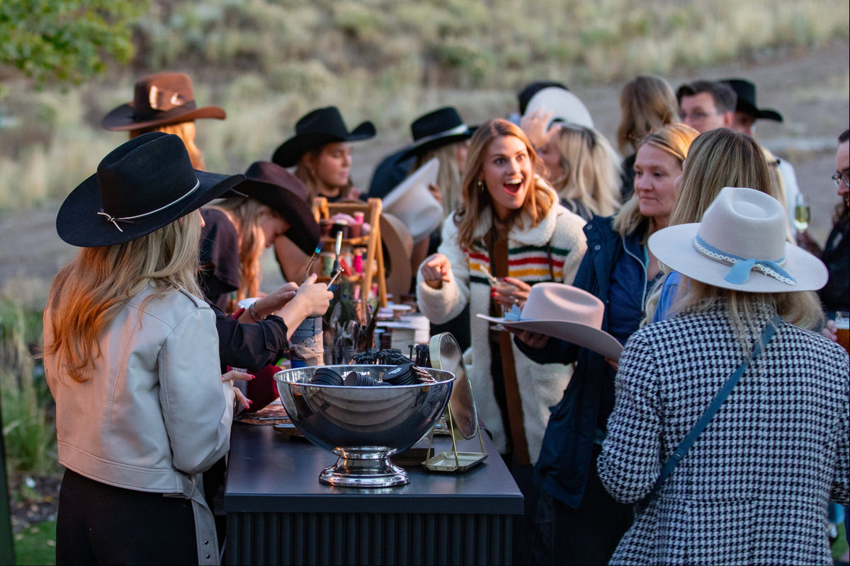 A group of people gathered outdoors at a social event or tailgate, standing around a table with drinks and a large metal bowl. Several people are wearing cowboy hats, and the background shows a dry grassy field with string lights overhead.