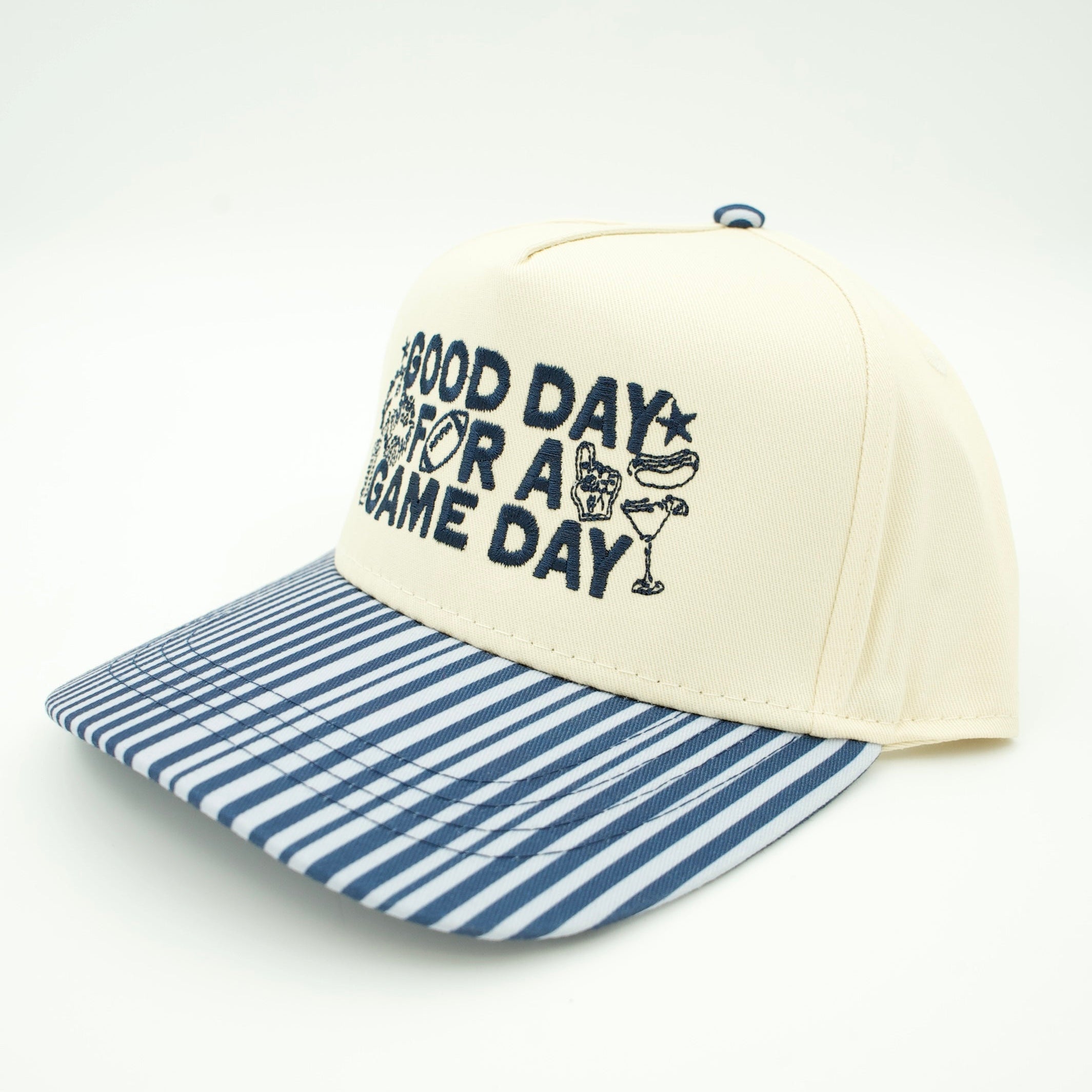 A baseball cap with an off-white crown, navy blue text reading 'GOOD DAY FOR A GAME DAY' with a cocktail graphic, and a blue and white striped visor, shown on a light gray background.