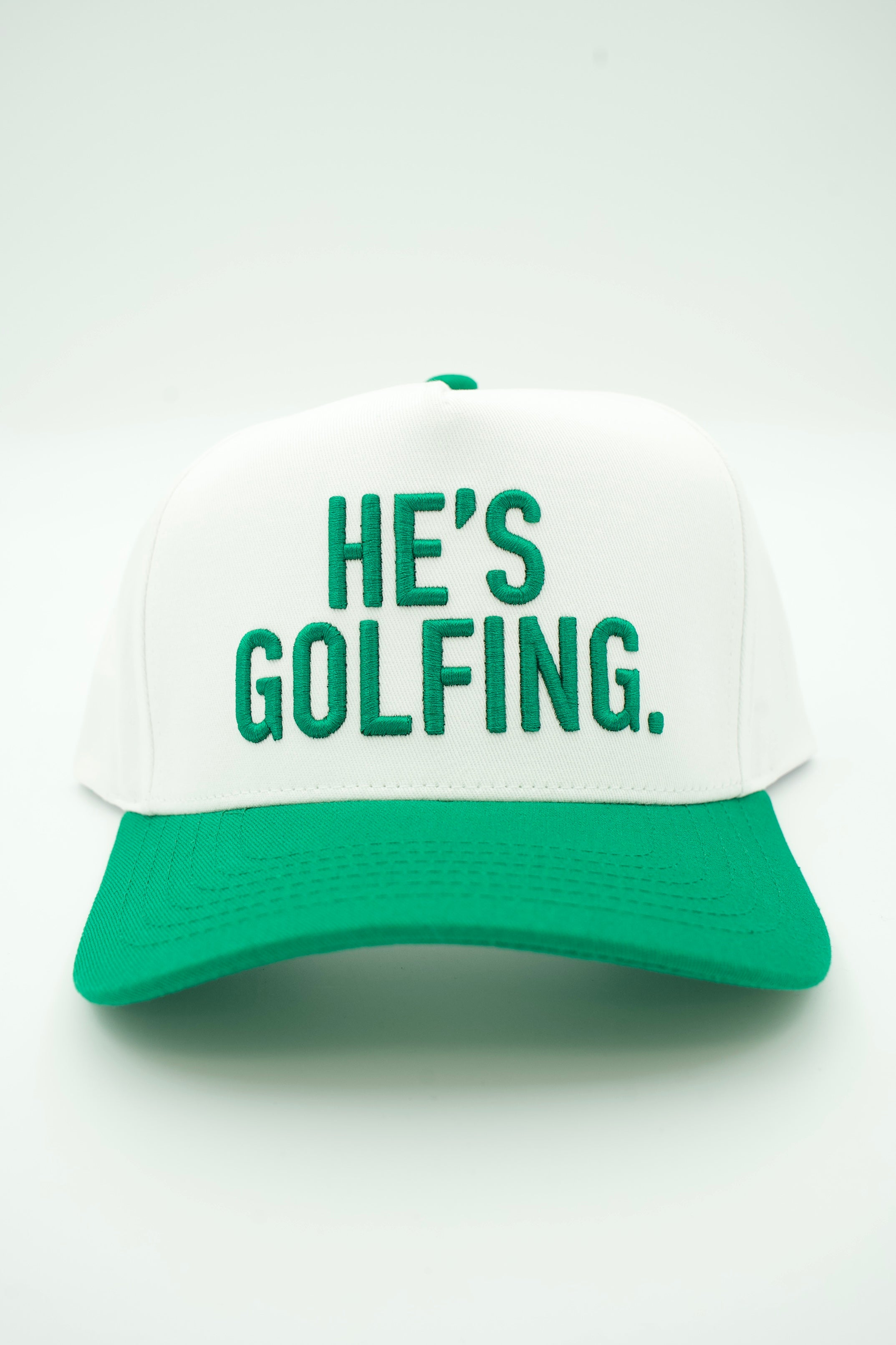 A white and green baseball cap with the phrase 'HE'S GOLFING.' embroidered in green on the front, set against a plain white background.