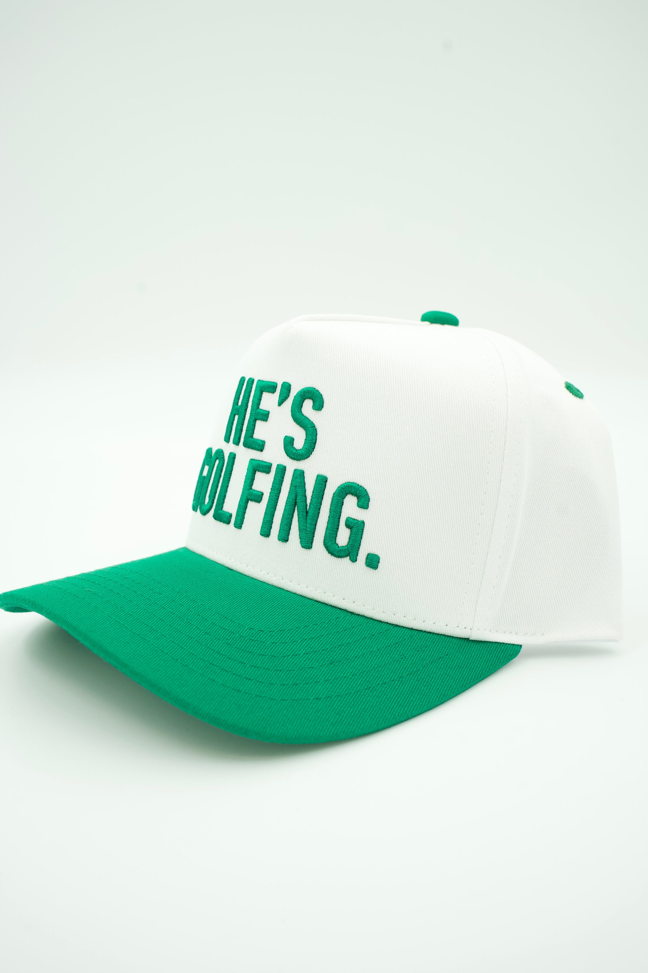 A white and green baseball cap with the text 'HE'S GOLFING.' embroidered in green on the front, set against a plain white background.
