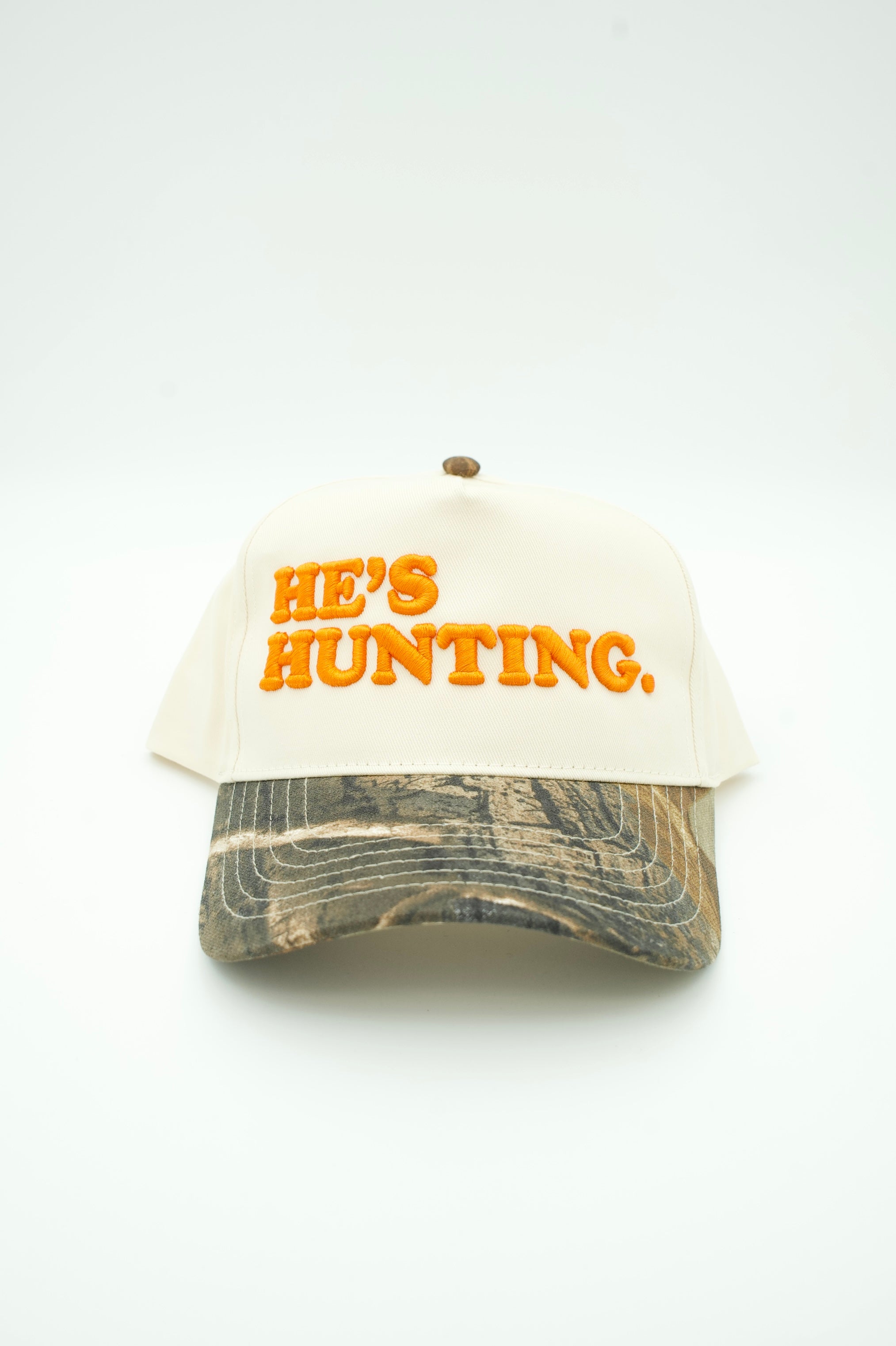 A trucker hat with an off-white front panel and mesh back, featuring the phrase 'HE'S HUNTING.' in orange embroidered letters.