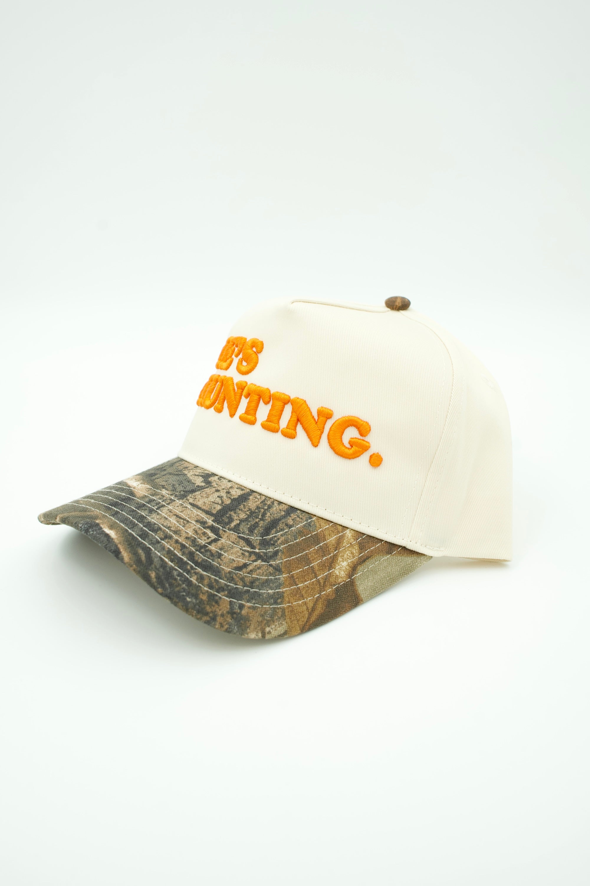 A baseball cap with an off-white crown, orange embroidered text reading 'IT'S HUNTING.', and a camouflage-patterned visor, shown on a plain light background.