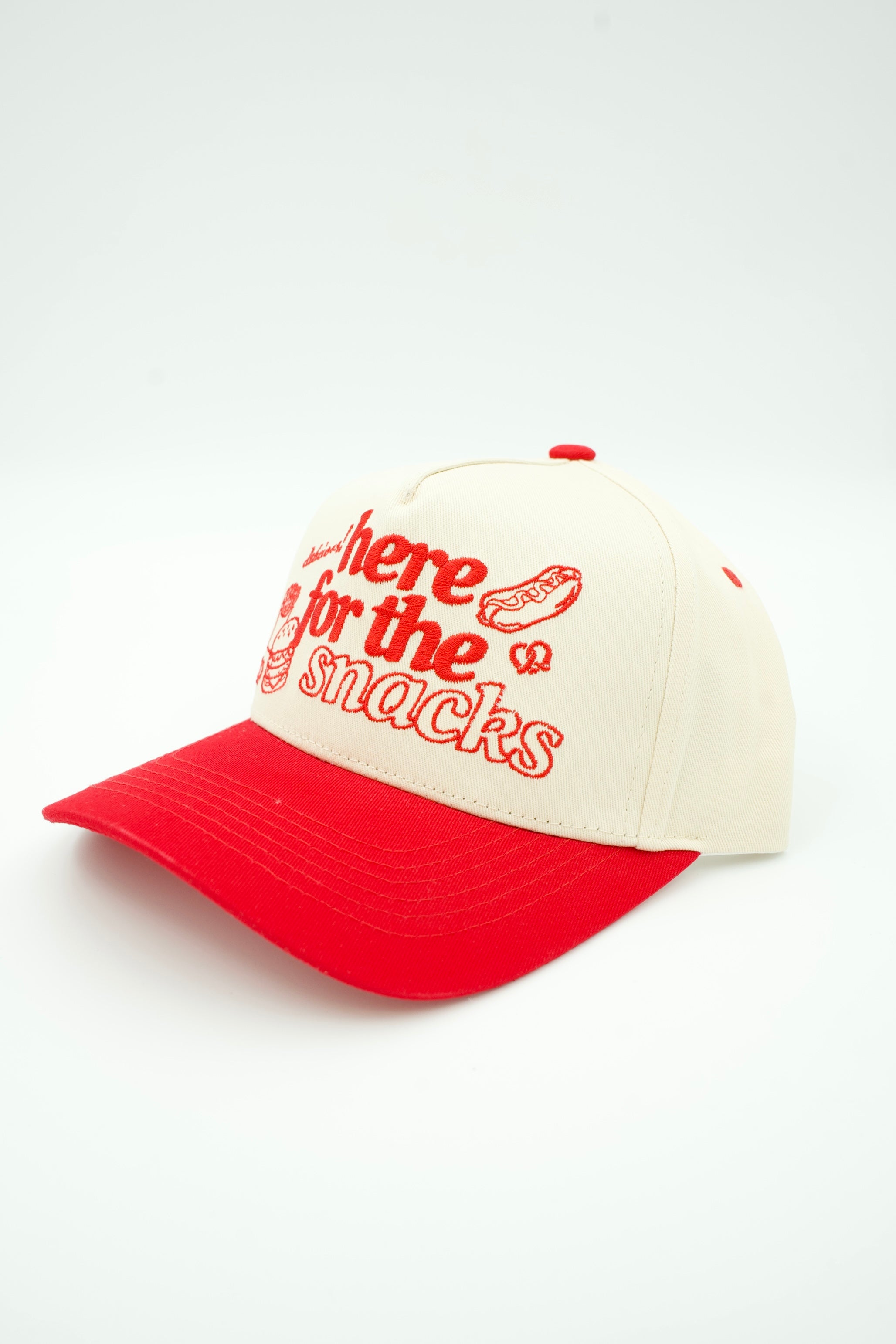 A two-tone baseball cap with a cream-colored crown and a bright red brim, featuring red embroidered text that reads 'Where the best snacks are' alongside a hot dog illustration.