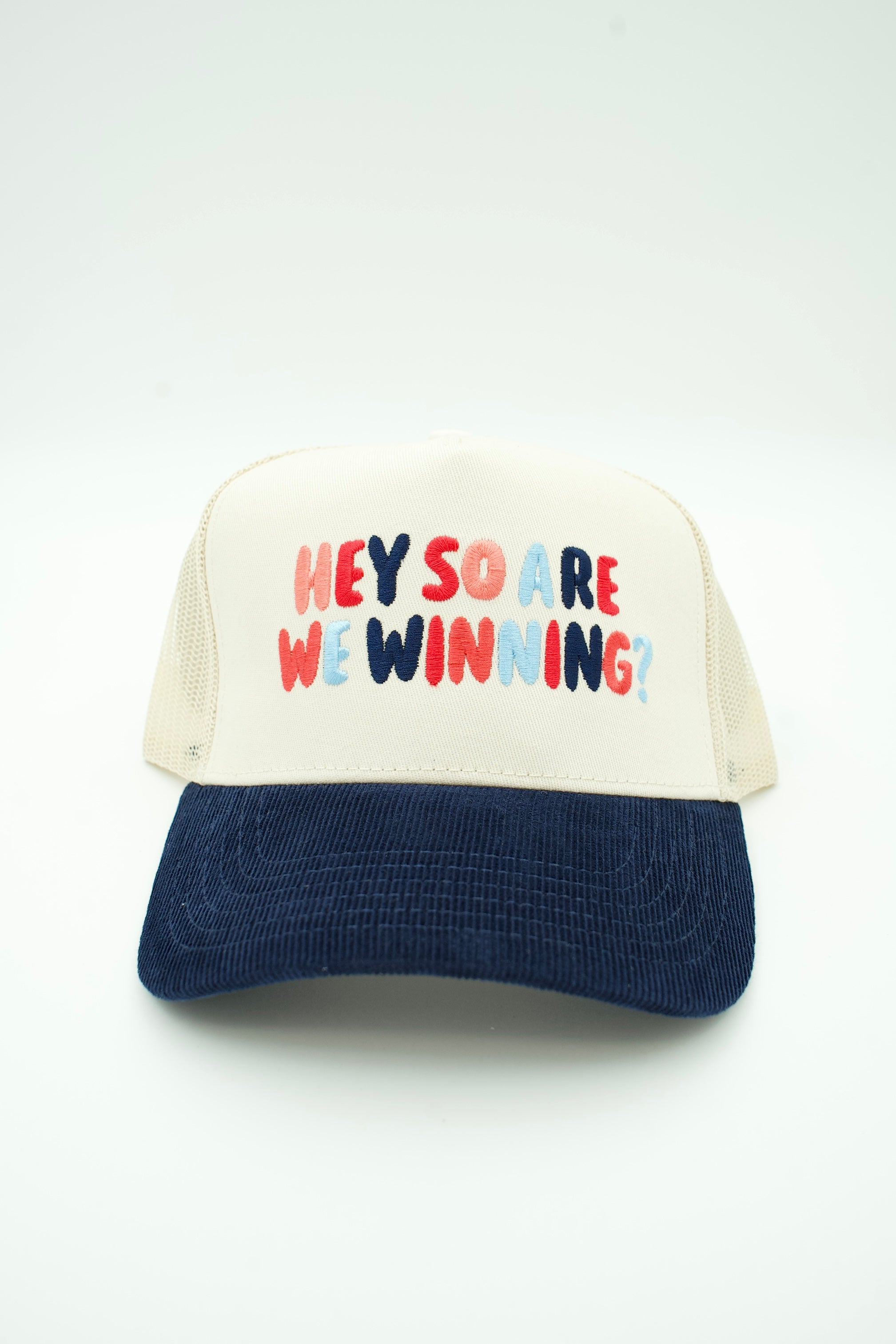 A trucker-style cap with an off-white front panel and mesh back, featuring the text 'HEY SO ARE WE WINNING?' in red, blue, and pink bubble letters, with a dark navy corduroy brim.