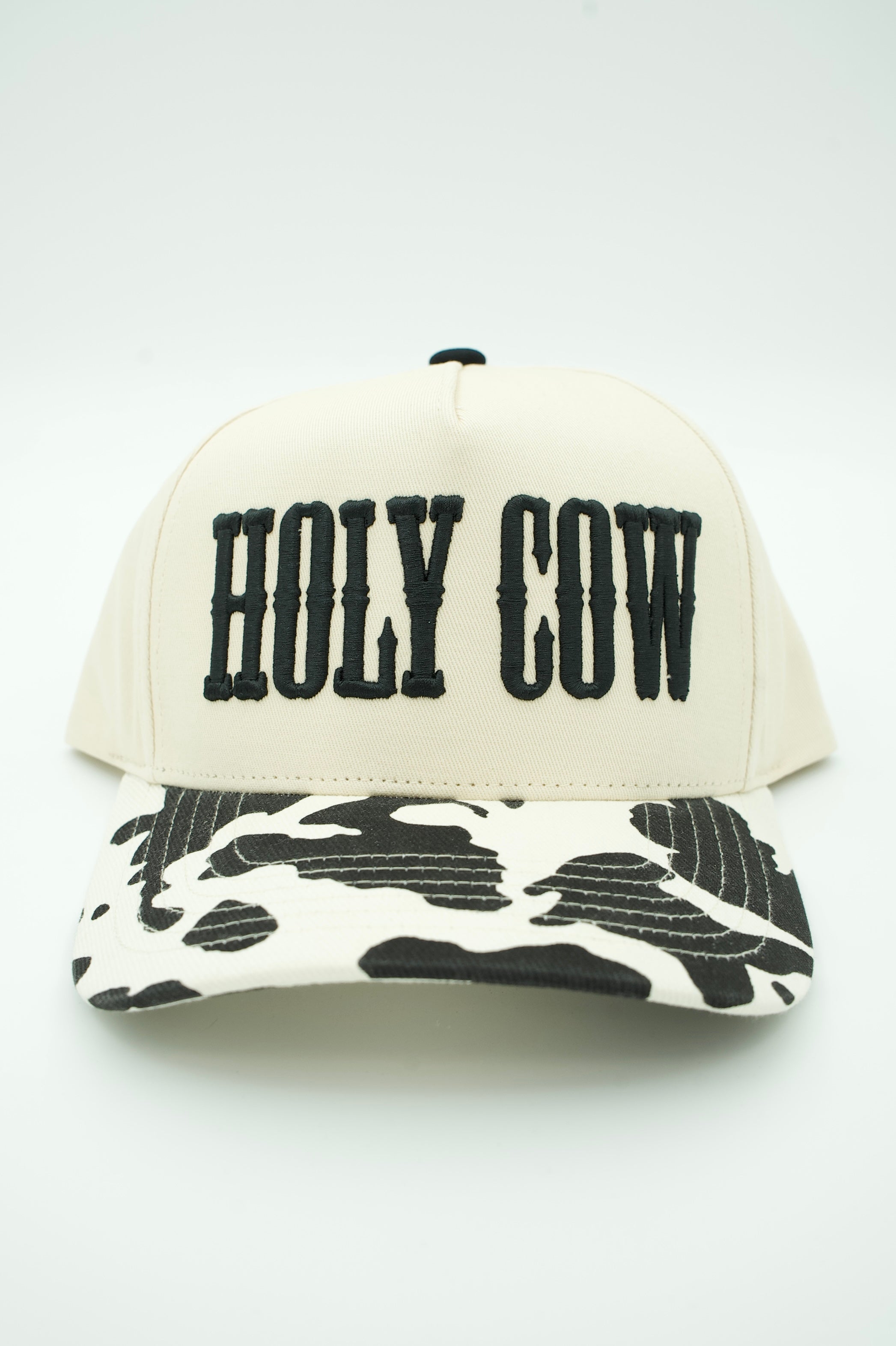 An off-white baseball cap with 'HOLY COW' embroidered in black block letters on the front and a black-and-white cow-print brim, shown against a plain white background.