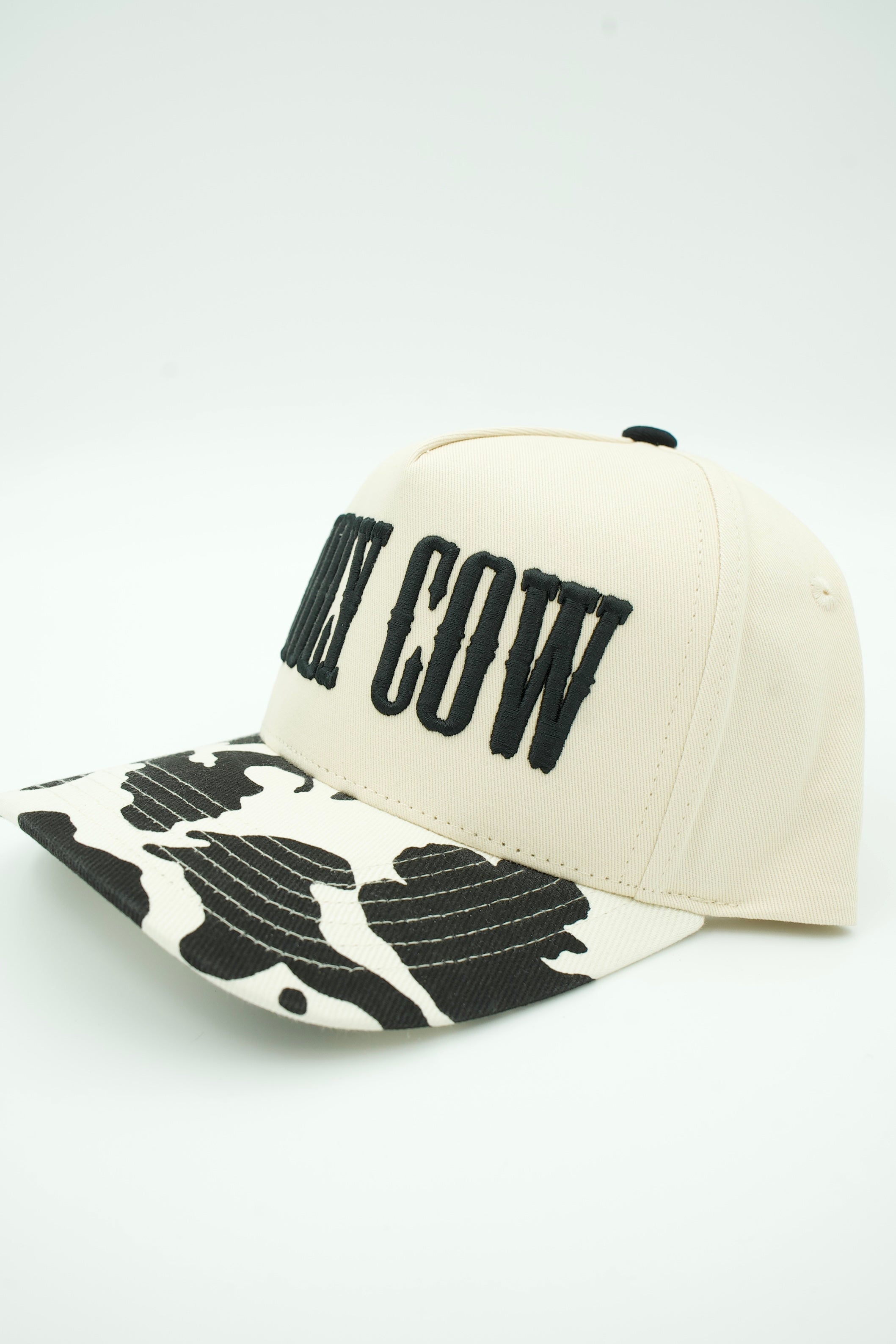 An off-white baseball cap with black embroidered text reading 'MY COW' on the front and a black-and-white cow-print pattern on the brim, shown on a light gray background.