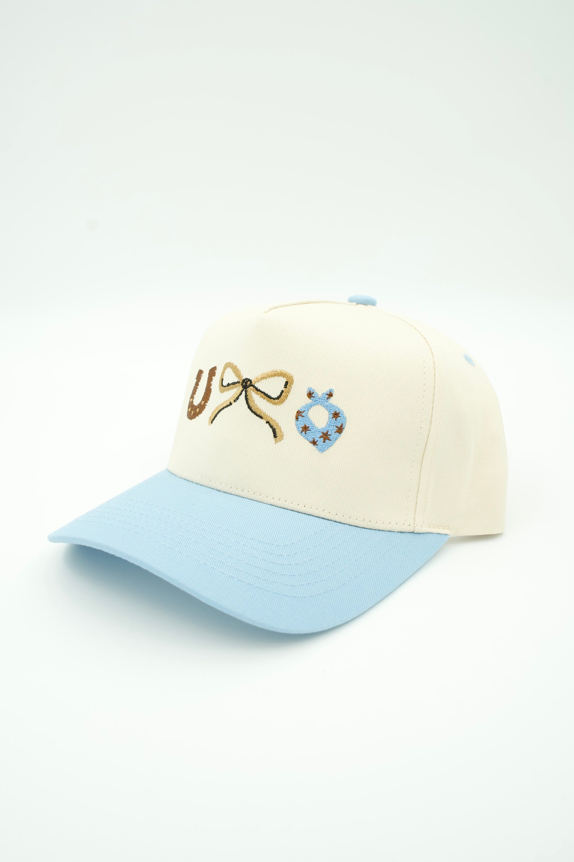A baseball cap with an off-white crown and a light blue brim, featuring embroidered details on the front including a gold bow and a blue bandana.