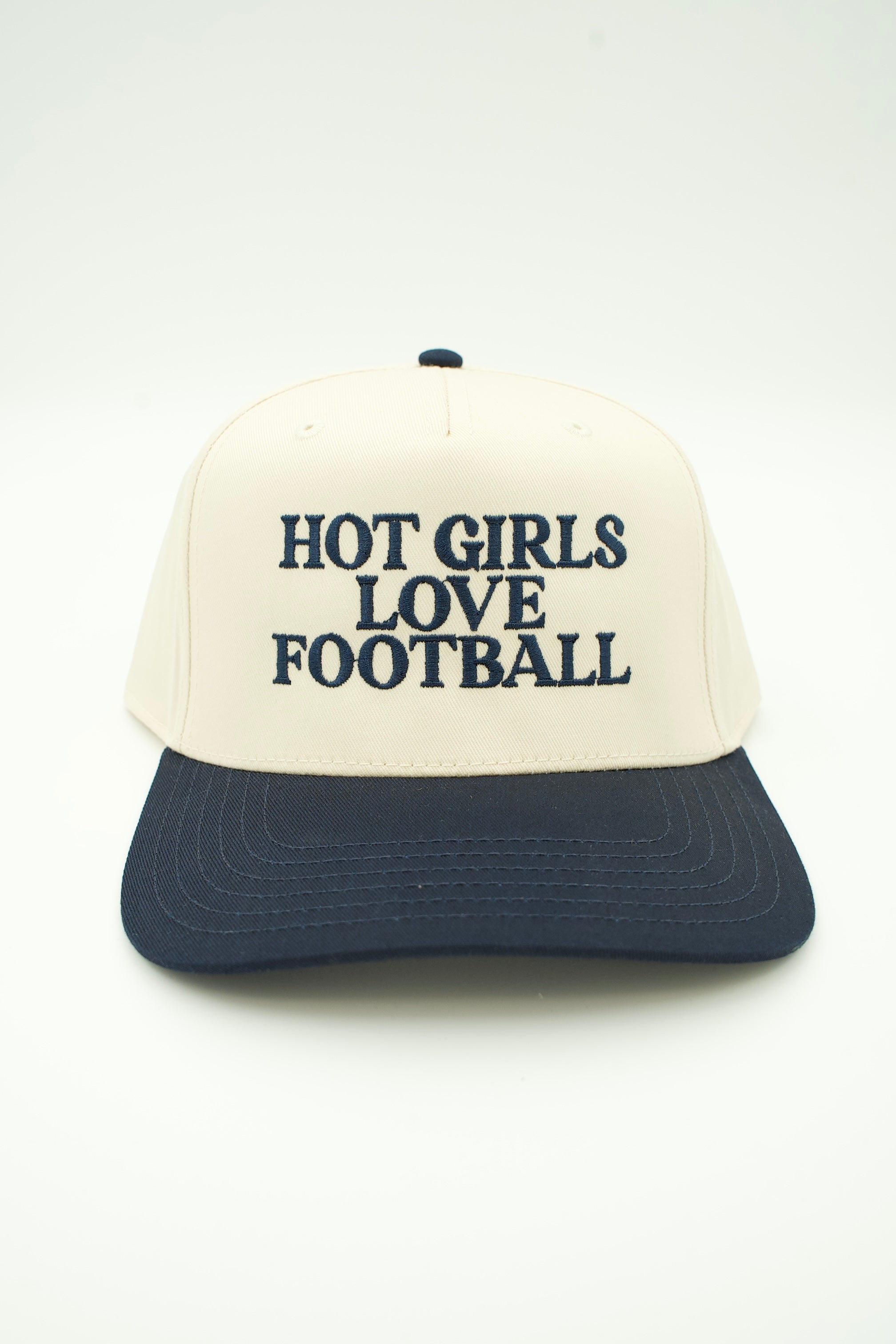 A two-tone baseball cap with an off-white crown and a navy blue brim, featuring the text 'HOT GIRLS LOVE FOOTBALL' embroidered in navy blue on the front. The cap is photographed against a clean, light off-white background.