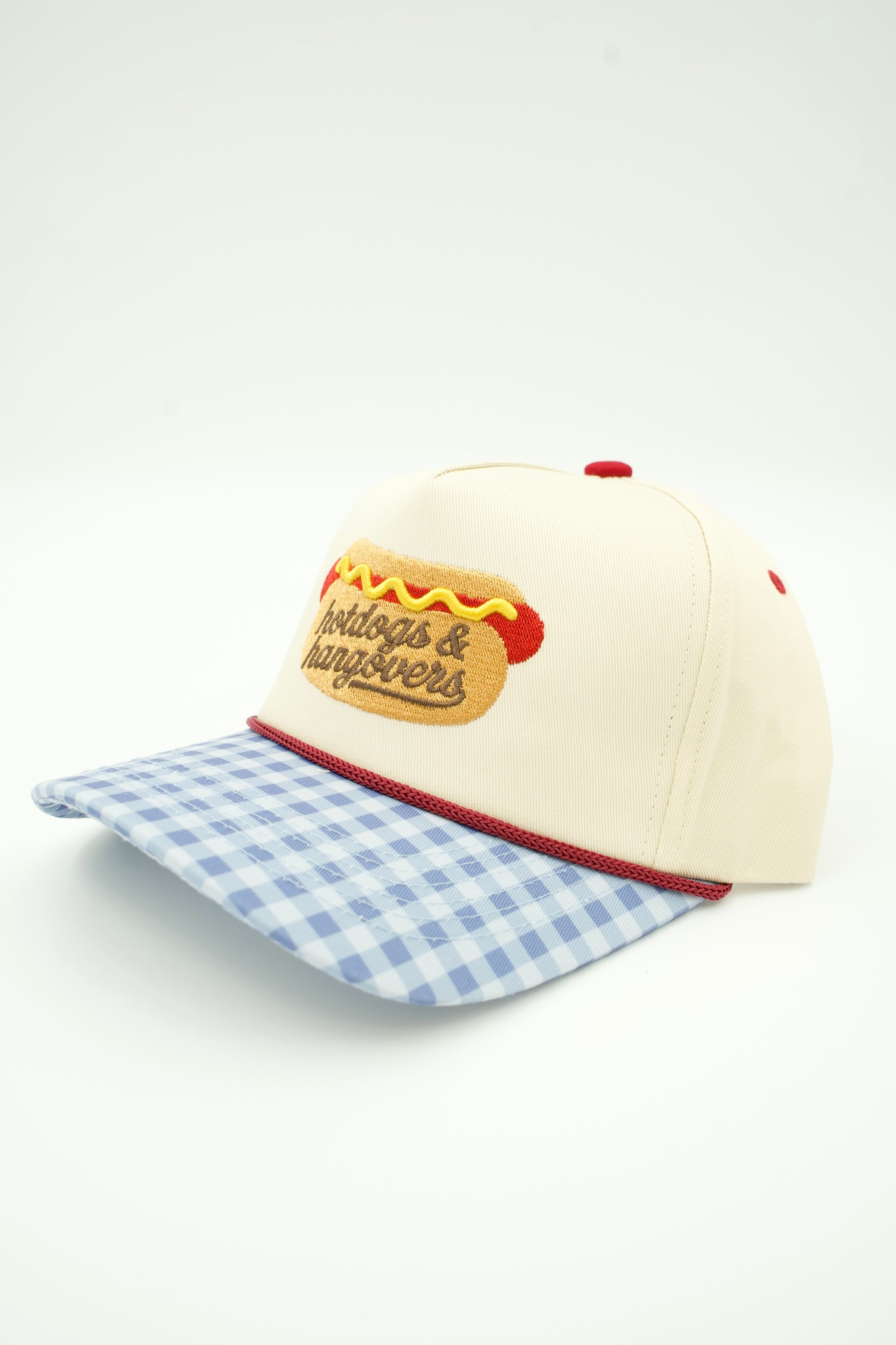 An off-white baseball cap with a blue and white gingham checkered brim, featuring an embroidered hot dog patch on the front that reads 'Hotdogs & Hangovers'.