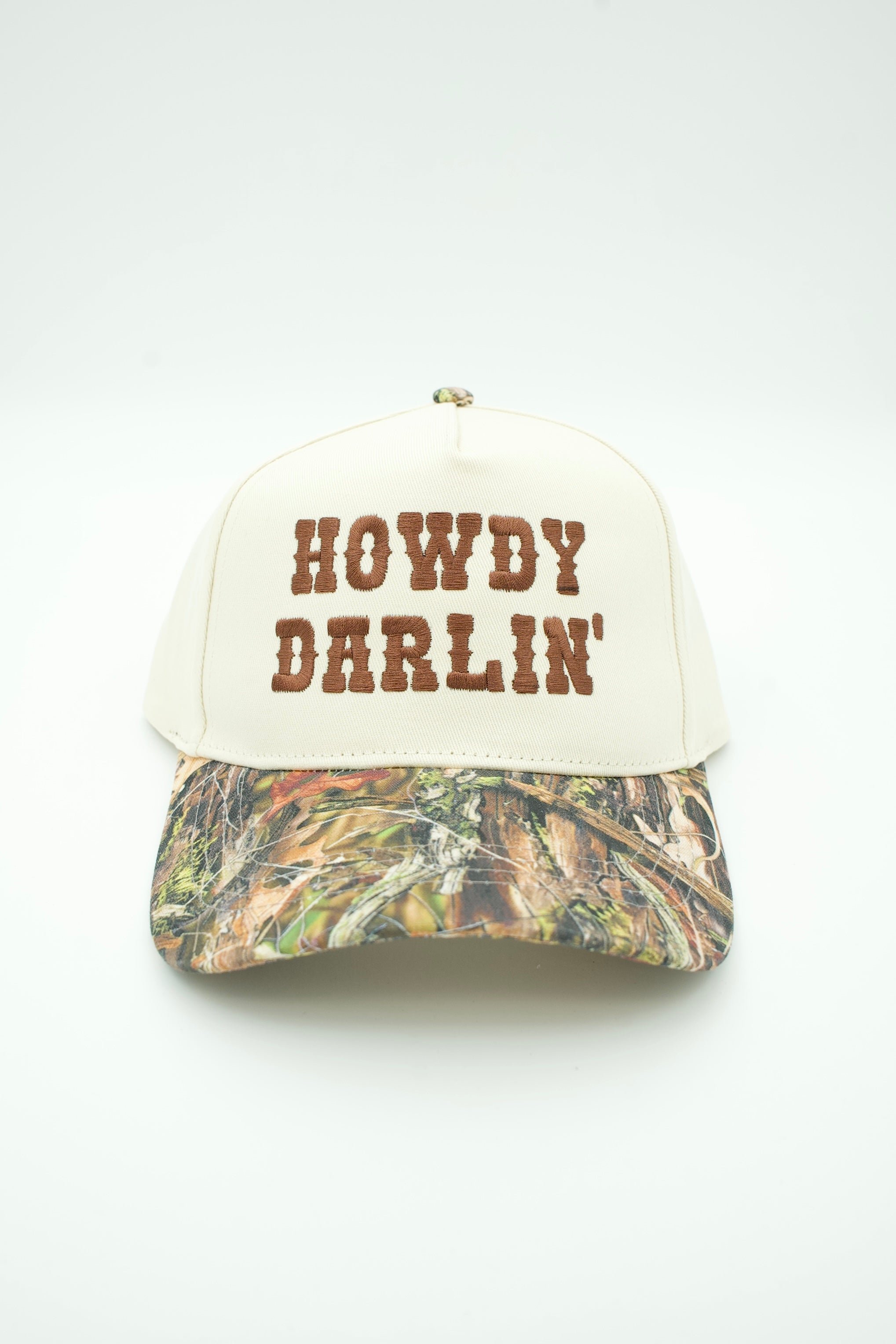 A baseball cap with an off-white front panel featuring the embroidered text 'HOWDY DARLIN'' in brown, and a camouflage-patterned visor in shades of green, brown, and beige.