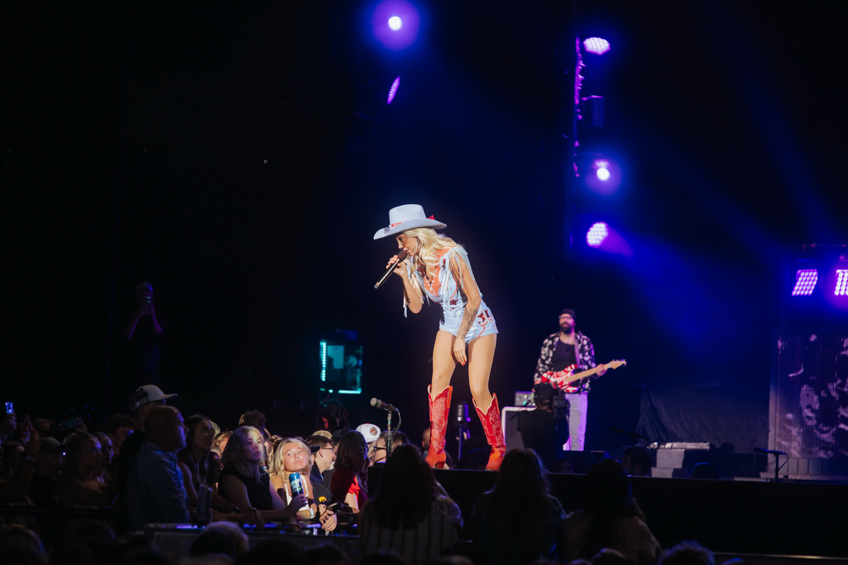 A live concert performance on a dark stage with blue and purple spotlights, featuring a female singer in a white cowboy hat, light blue outfit, and red boots holding a microphone, with a guitarist and band members in the background.