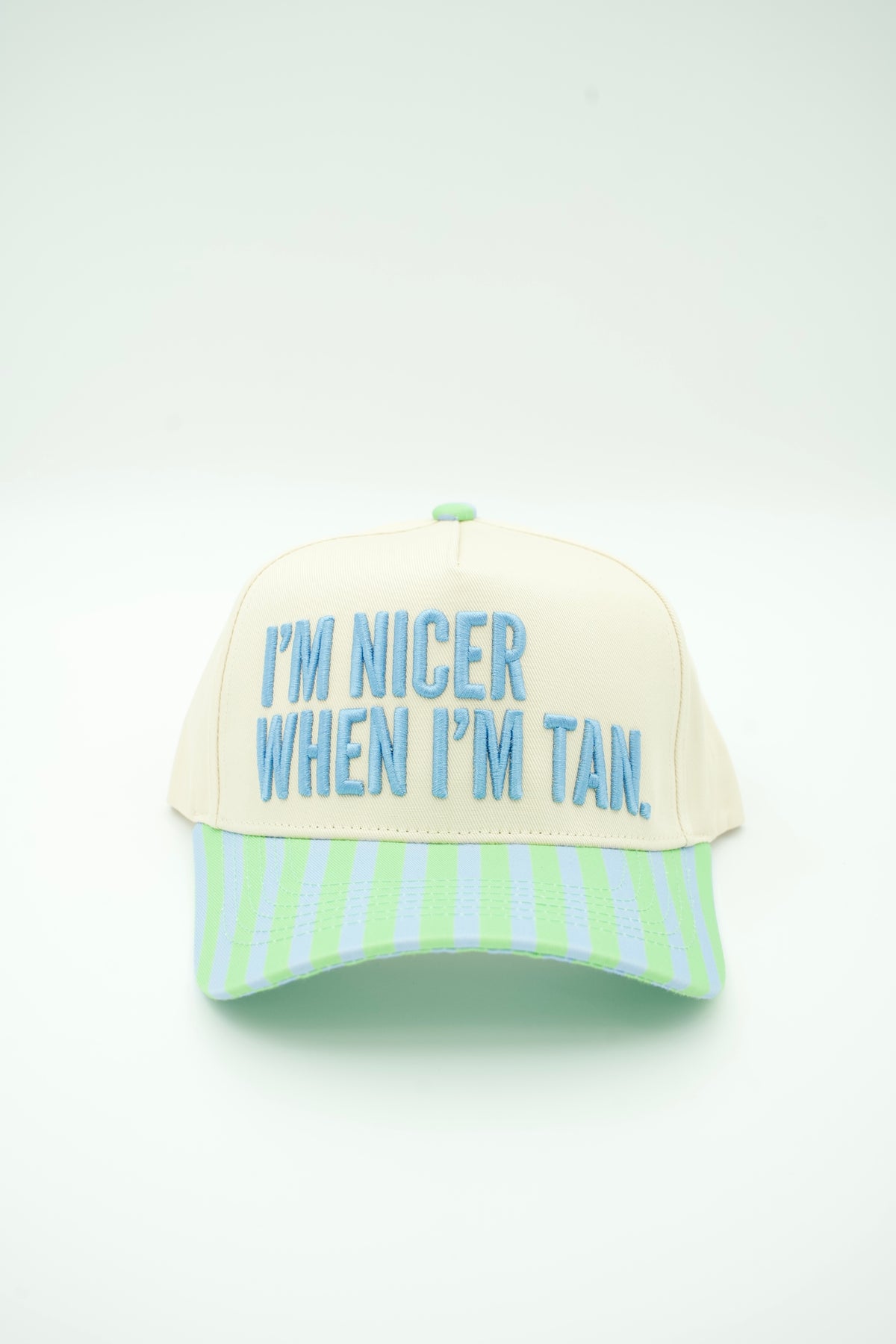 Front image of a cream canvas trucker hat with "I'M NICER WHEN I'M TAN" embroidered on it with a baby blue and mint green striped bill