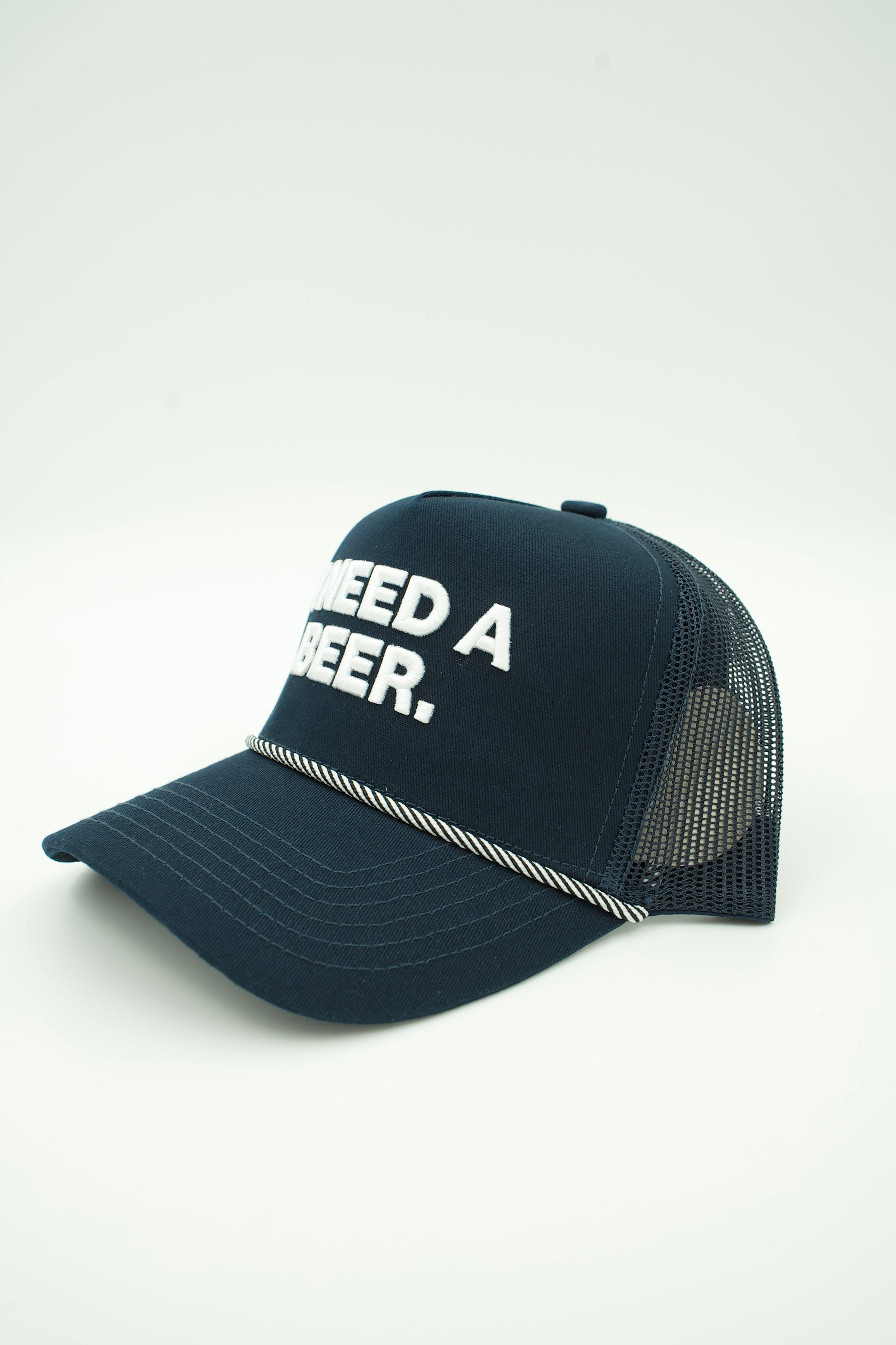 A navy blue trucker hat with white embroidered text reading 'NEED A BEER.' on the front panel and a thin white rope detail across the brim, shown on a light gray background.