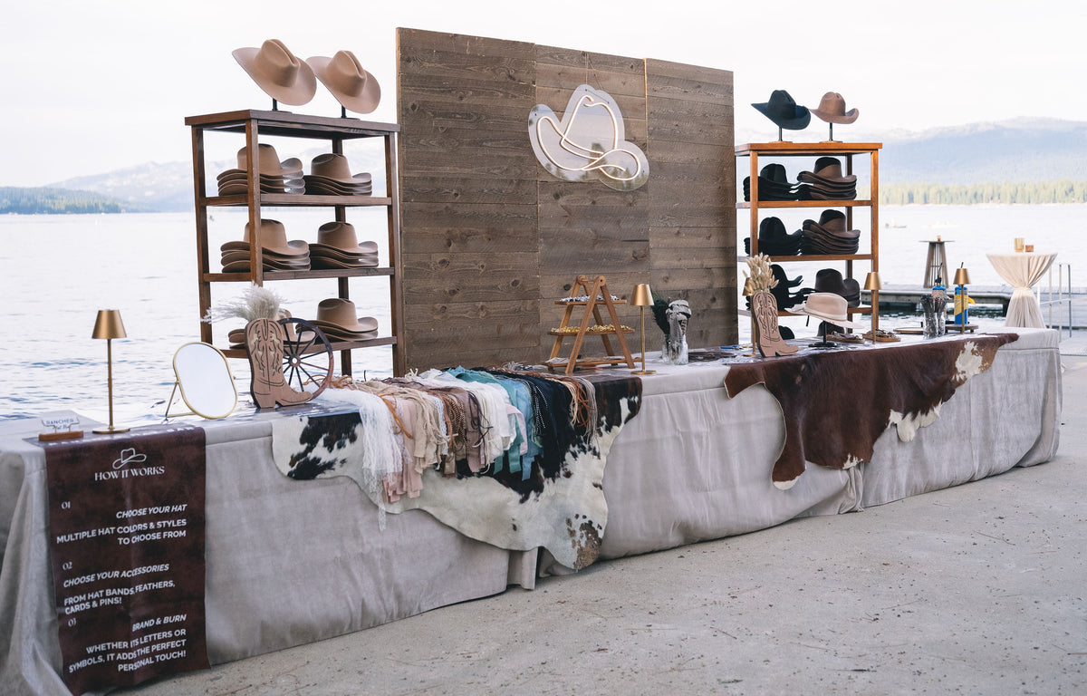 A styled outdoor display table featuring a collection of cowboy hats, leather aprons, cowhide rugs, and rustic decor, set against a backdrop of a lake and mountains. A wooden sign on the left reads 'HARVEY BARRON'.