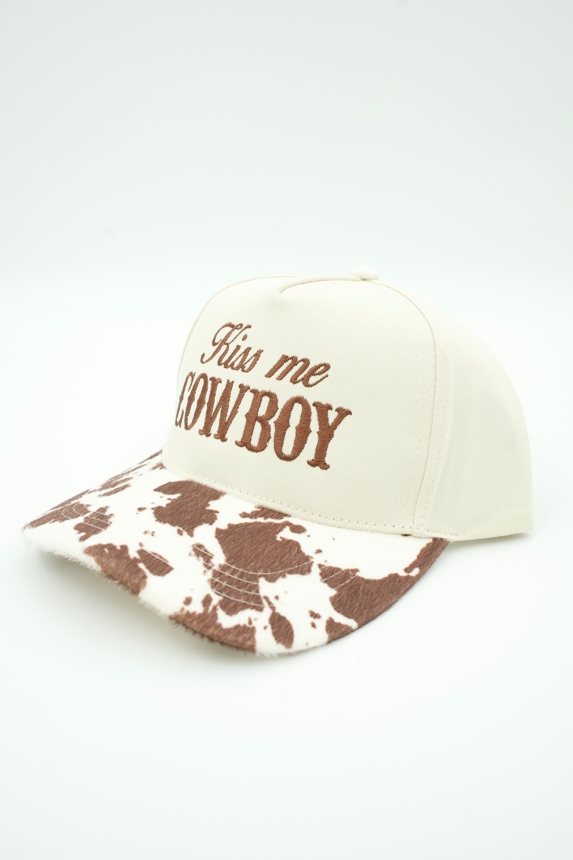 An off-white baseball cap with brown embroidered text reading 'Kiss me COWBOY' on the front, and a brown-and-white cow-print patterned brim.