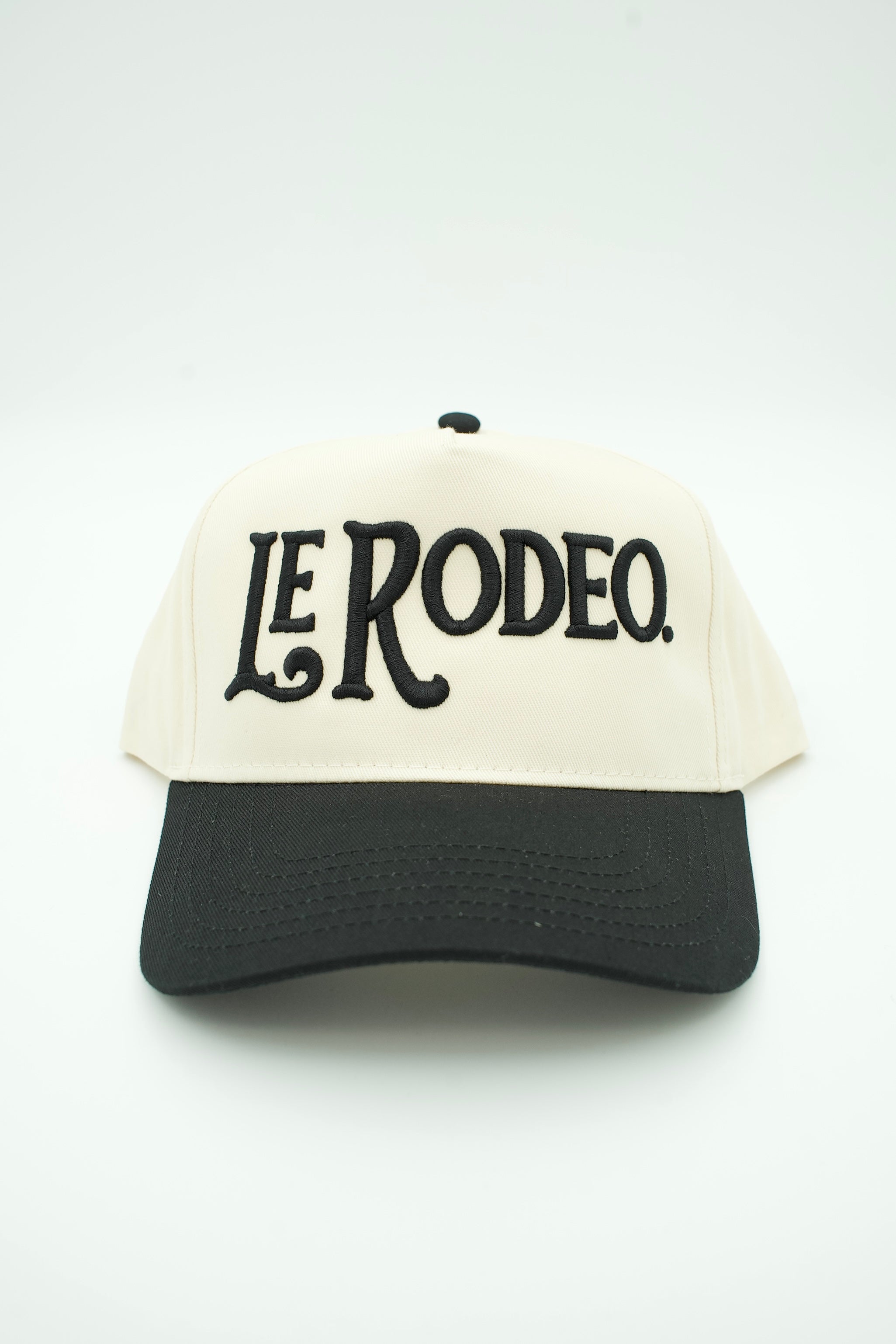 A two-tone trucker hat with an off-white front panel and brim, and black mesh side/back panels. The front features the text 'LE RODEO.