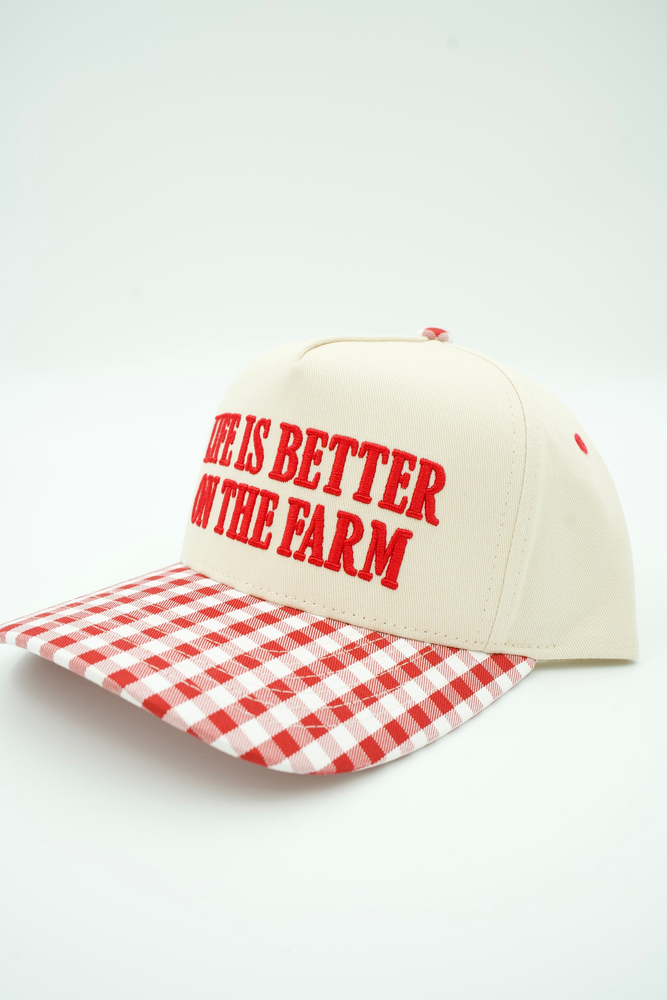 An off-white trucker hat with red text reading 'LIFE IS BETTER ON THE FARM' and a red-and-white gingham patterned brim, photographed on a clean white background.