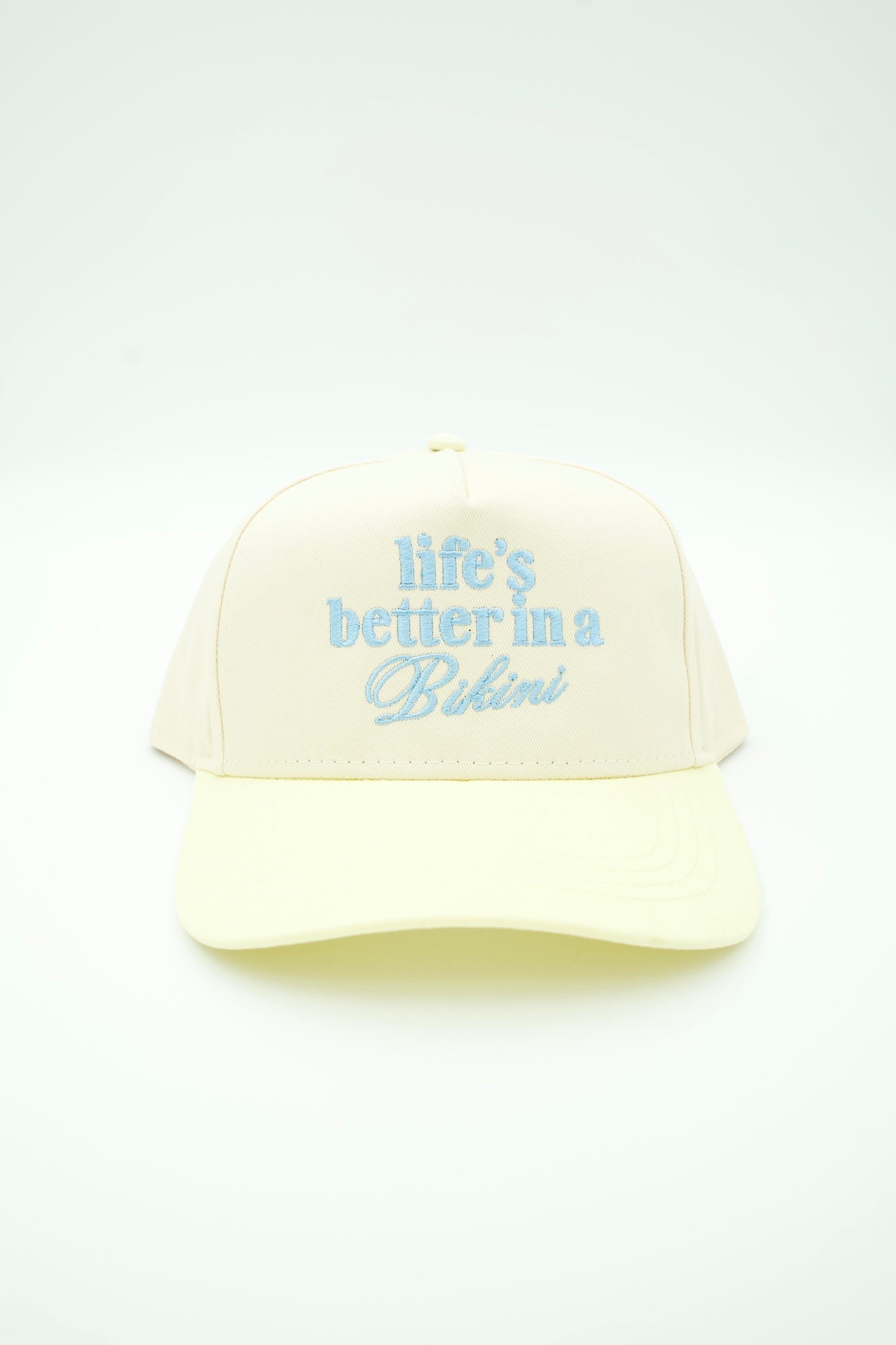 A cream-colored trucker-style cap with light blue embroidered text reading 'life's better in a Bikini' on the front, set against a plain white background.