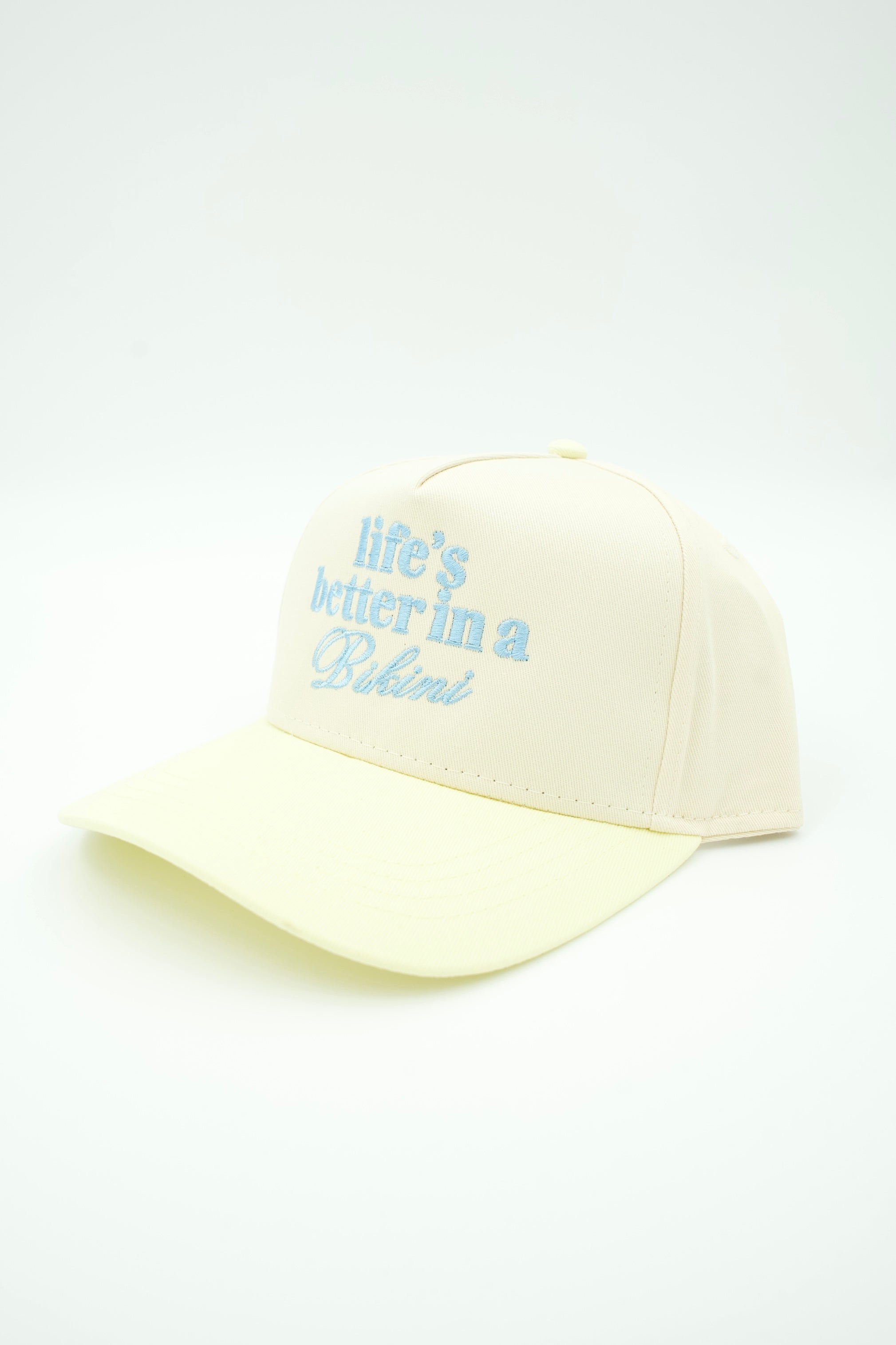 A pale cream baseball cap with light blue embroidered text reading 'life's better in a Bahini' on the front, shown on a clean white background.
