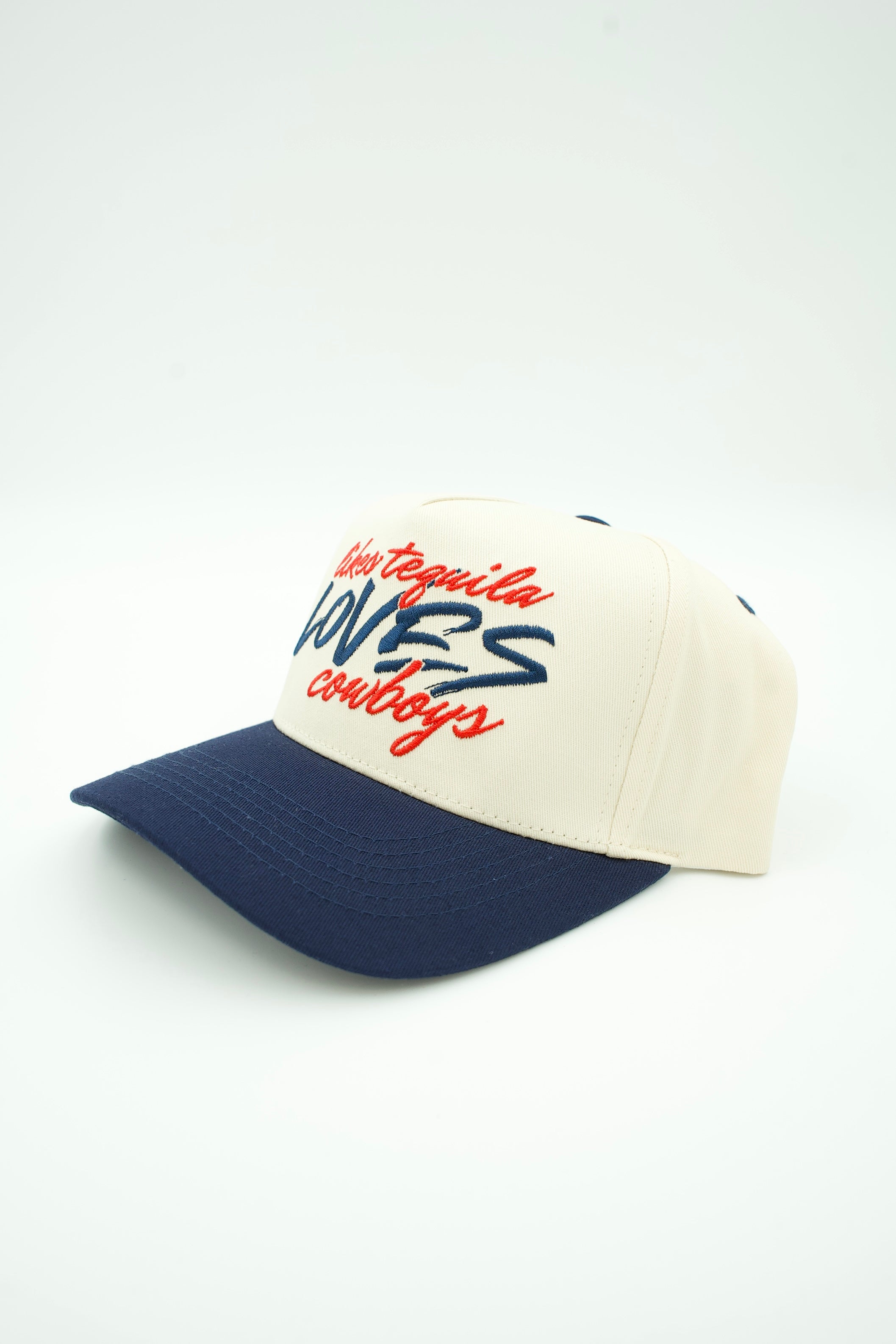 A baseball cap with an off-white crown and a navy blue curved brim, featuring embroidered text on the front that reads 'Tequila Lovers Cowboys' in red and navy blue.