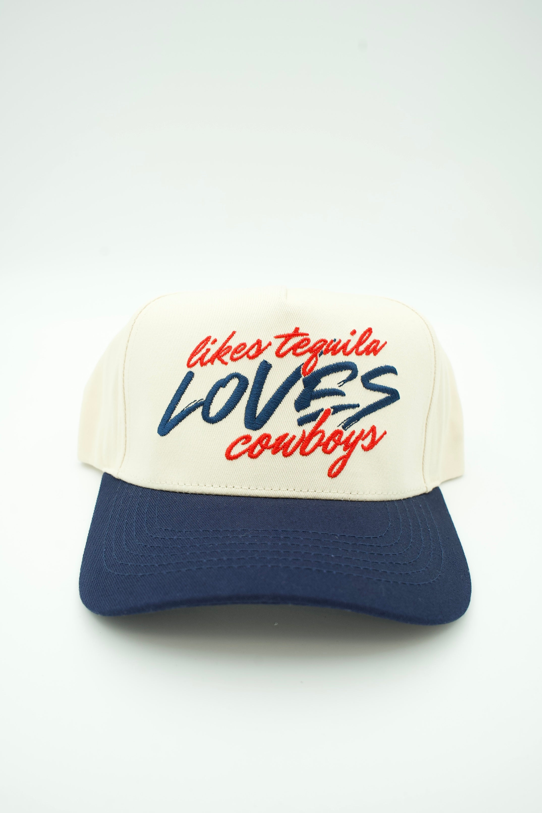 A two-tone trucker cap with an off-white front panel and brim, and a navy blue curved brim. The front panel features embroidered text reading 'likes tequila LOVES cowboys' in red and navy blue.