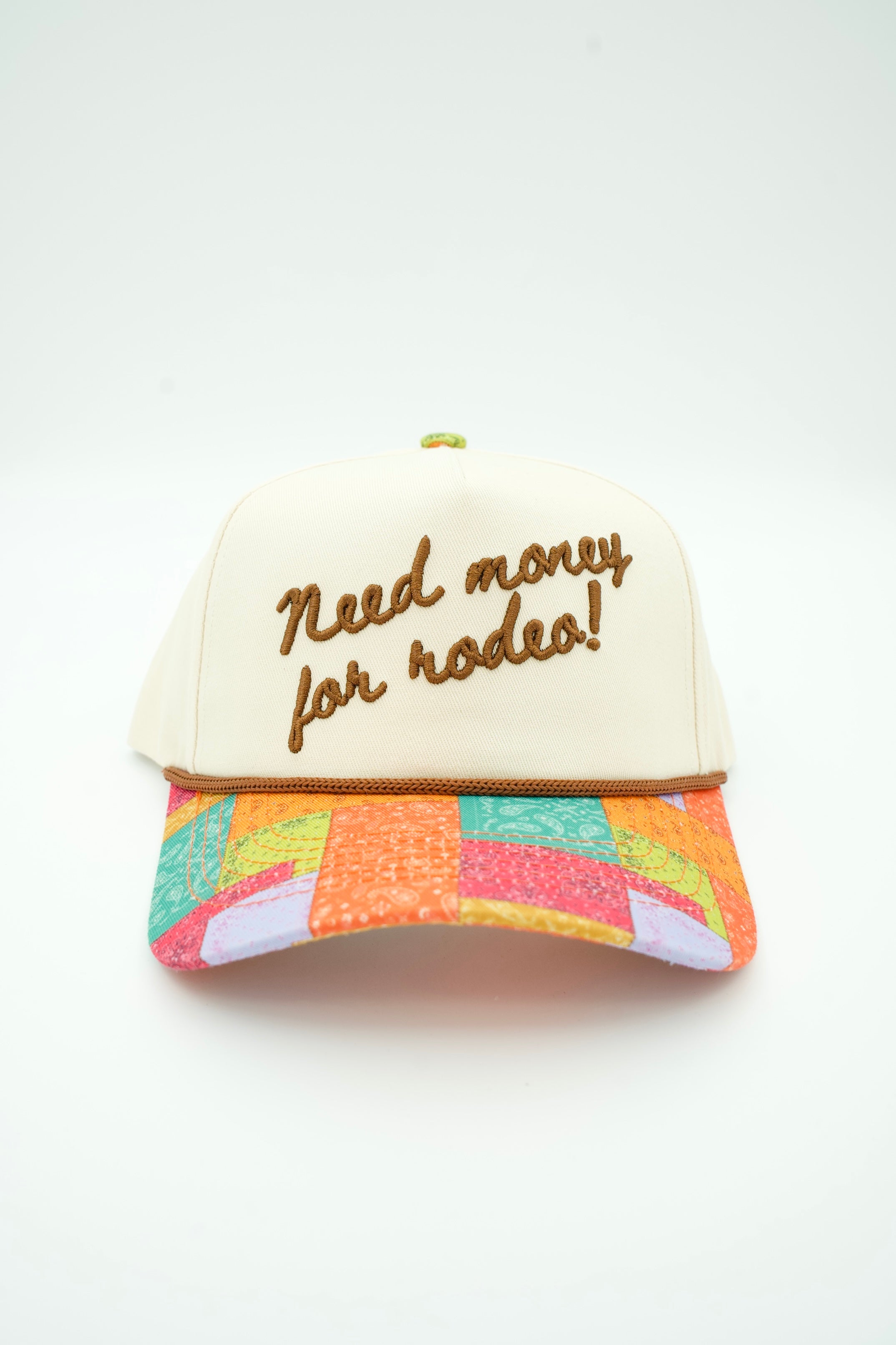 A cream-colored trucker-style cap with brown embroidered text reading 'Need money for rodeo!' on the front. The brim features a colorful patchwork pattern in orange, pink, green, and yellow.