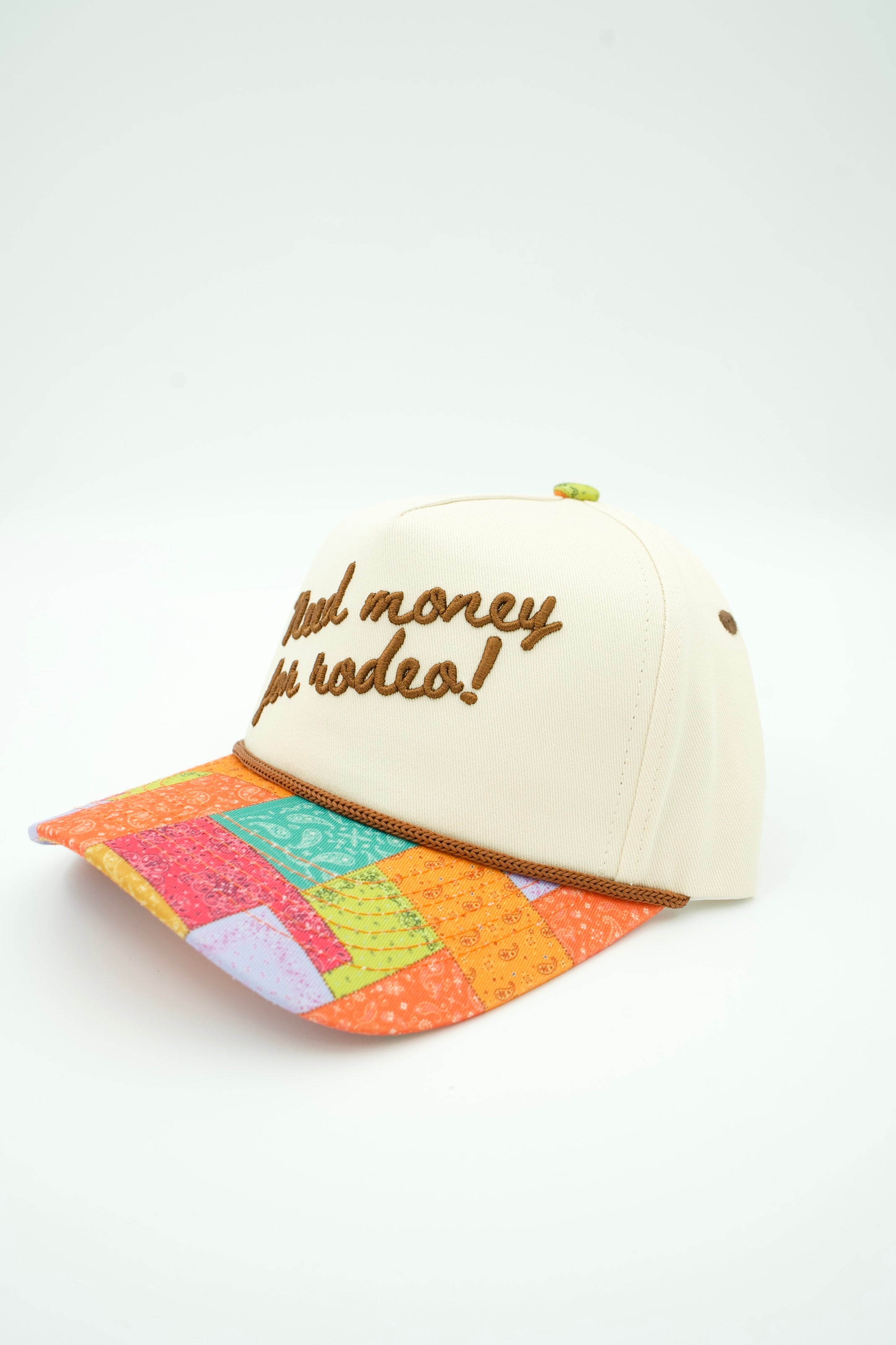 An off-white baseball cap with brown embroidered text reading 'feel money free! rodeo!' on the front, featuring a colorful patchwork-patterned brim and a small green button on top, set against a plain white background.