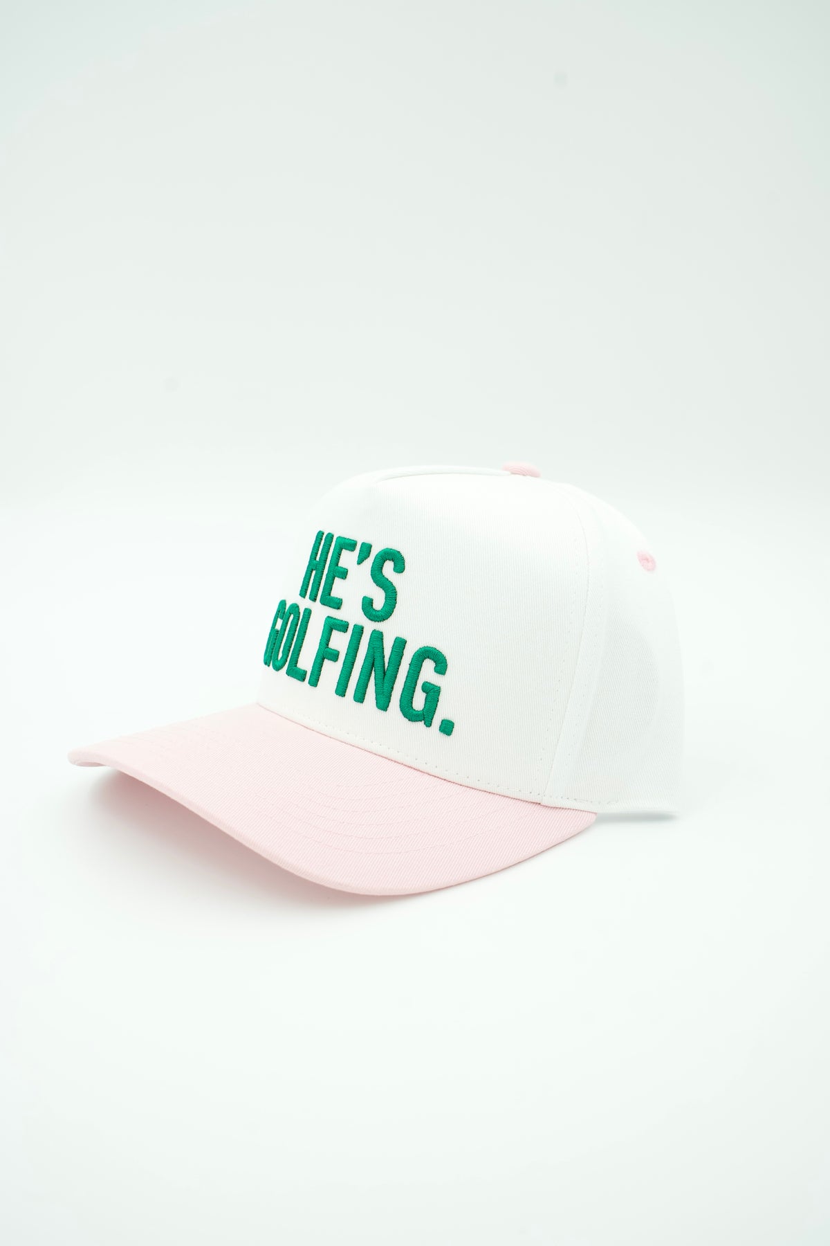 A two-tone baseball cap with a white crown, light pink brim, and green embroidered text reading 'HE'S GOLFING.' on the front, set against a plain white background.