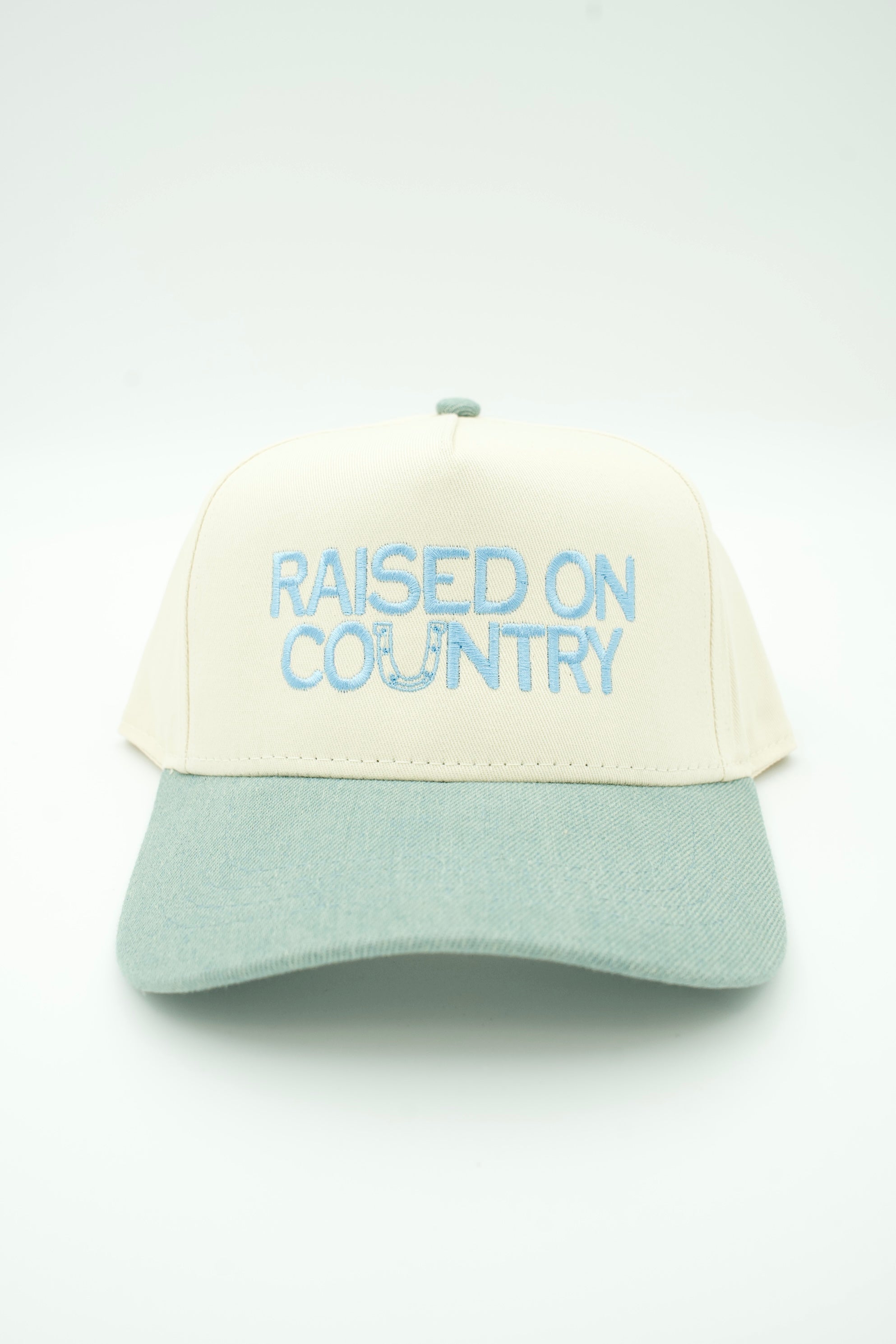A two-tone baseball cap with an off-white crown and a denim curved brim, featuring the phrase 'RAISED ON COUNTRY' embroidered in light blue on the front.