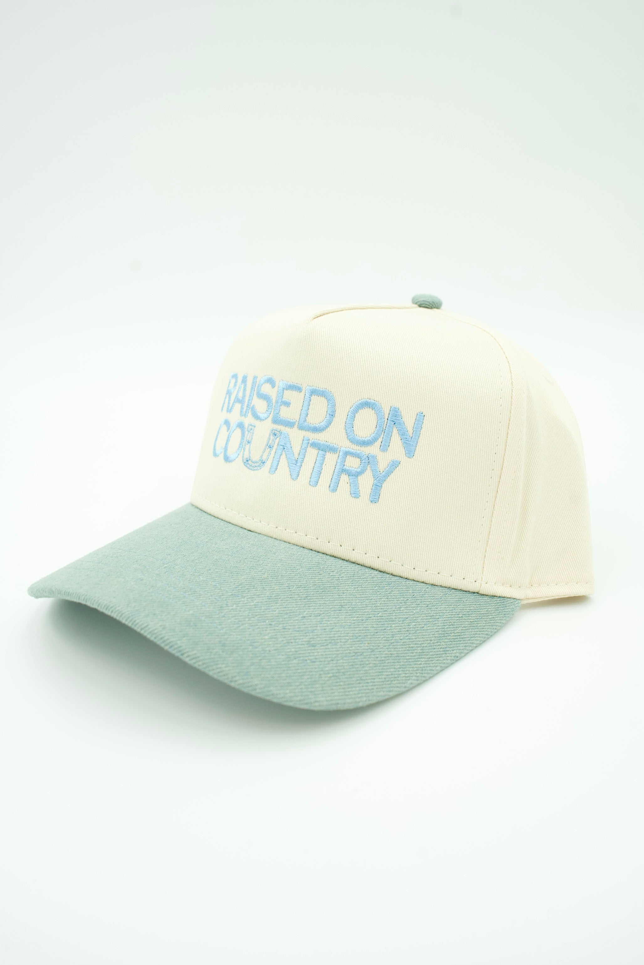 A two-tone baseball cap with an off-white crown and a sage green brim, featuring the embroidered text 'RAISED ON COUNTRY' in light blue on the front.