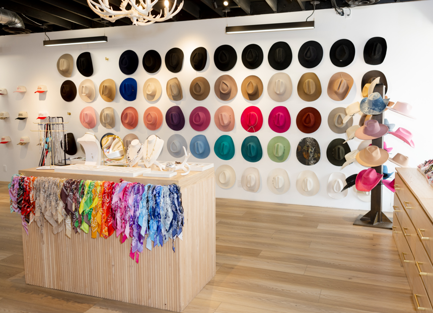 A retail display showcasing a wide collection of wide-brimmed hats. The wall behind the products features rows of colorful, neutral, and dark wide-brimmed hats, while a light wood counter in the foreground is draped with vibrant, multicolored fringed scarves.