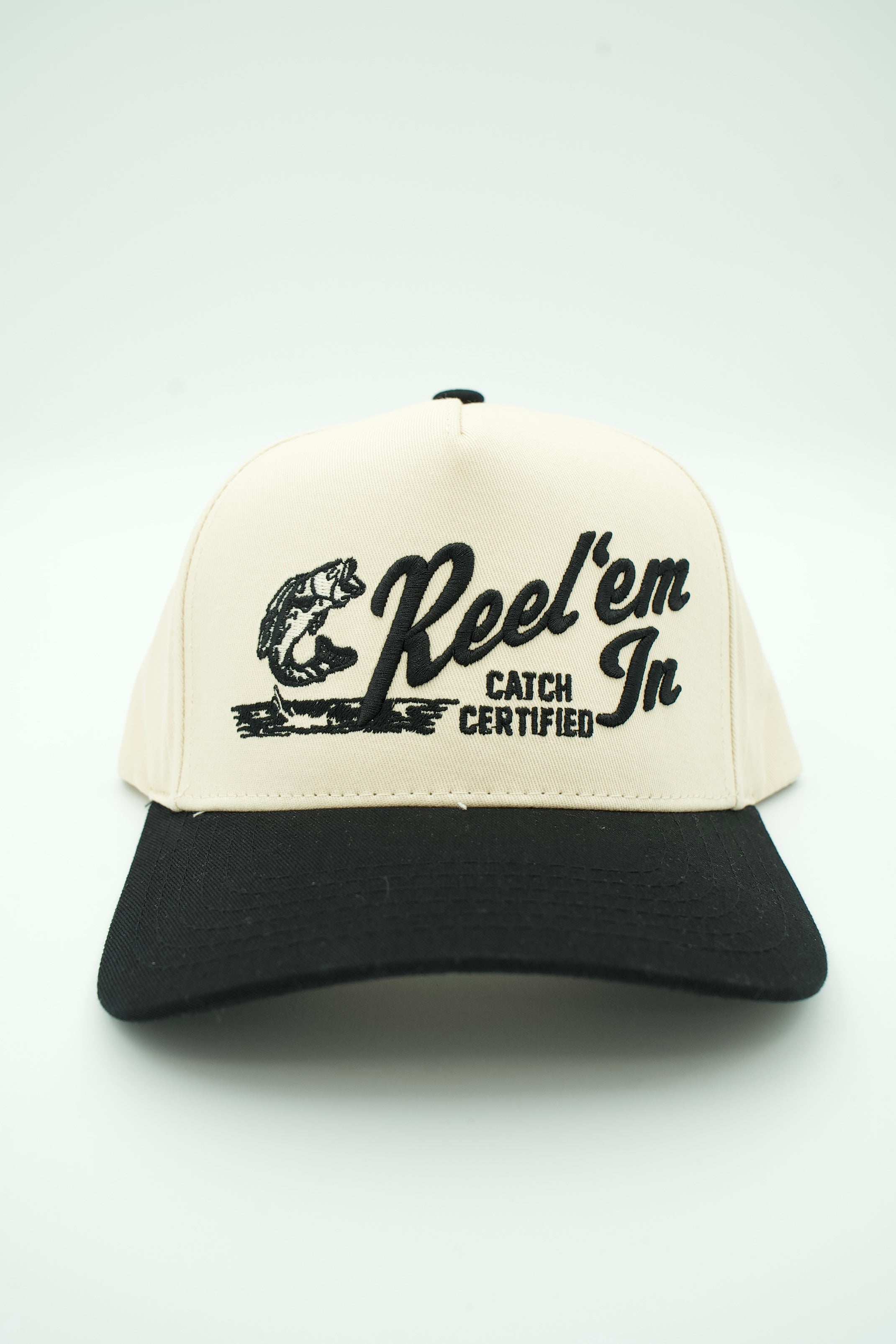 A two-tone trucker-style baseball cap with an off-white front panel and black brim, featuring a black graphic of a fish and the text 'Reel'em In' and 'CATCH CERTIFIED' on the front.