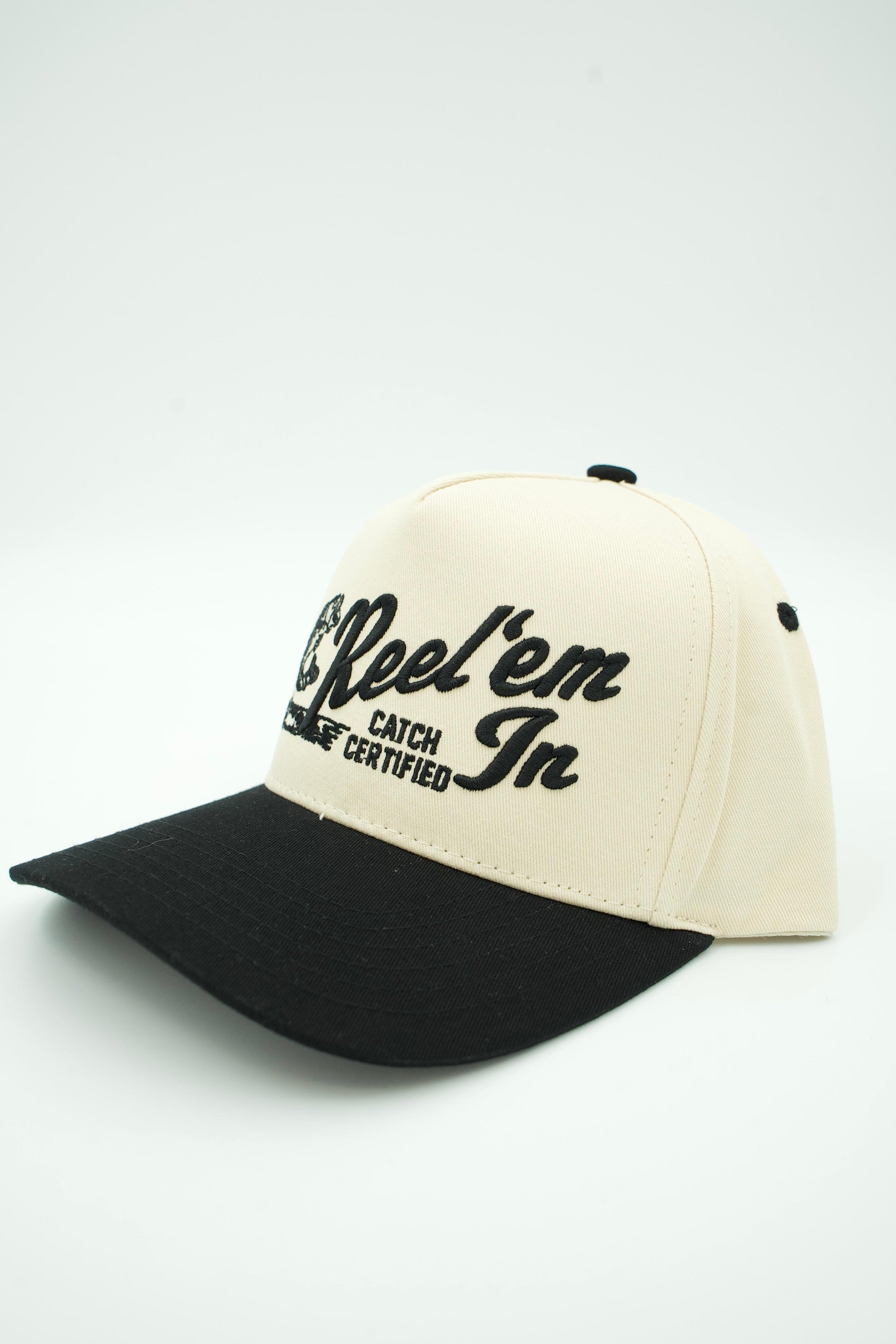 A two-tone baseball cap with an off-white crown and a black curved brim, featuring black embroidered text on the front that reads 'Reel 'em Catch Certified In'.