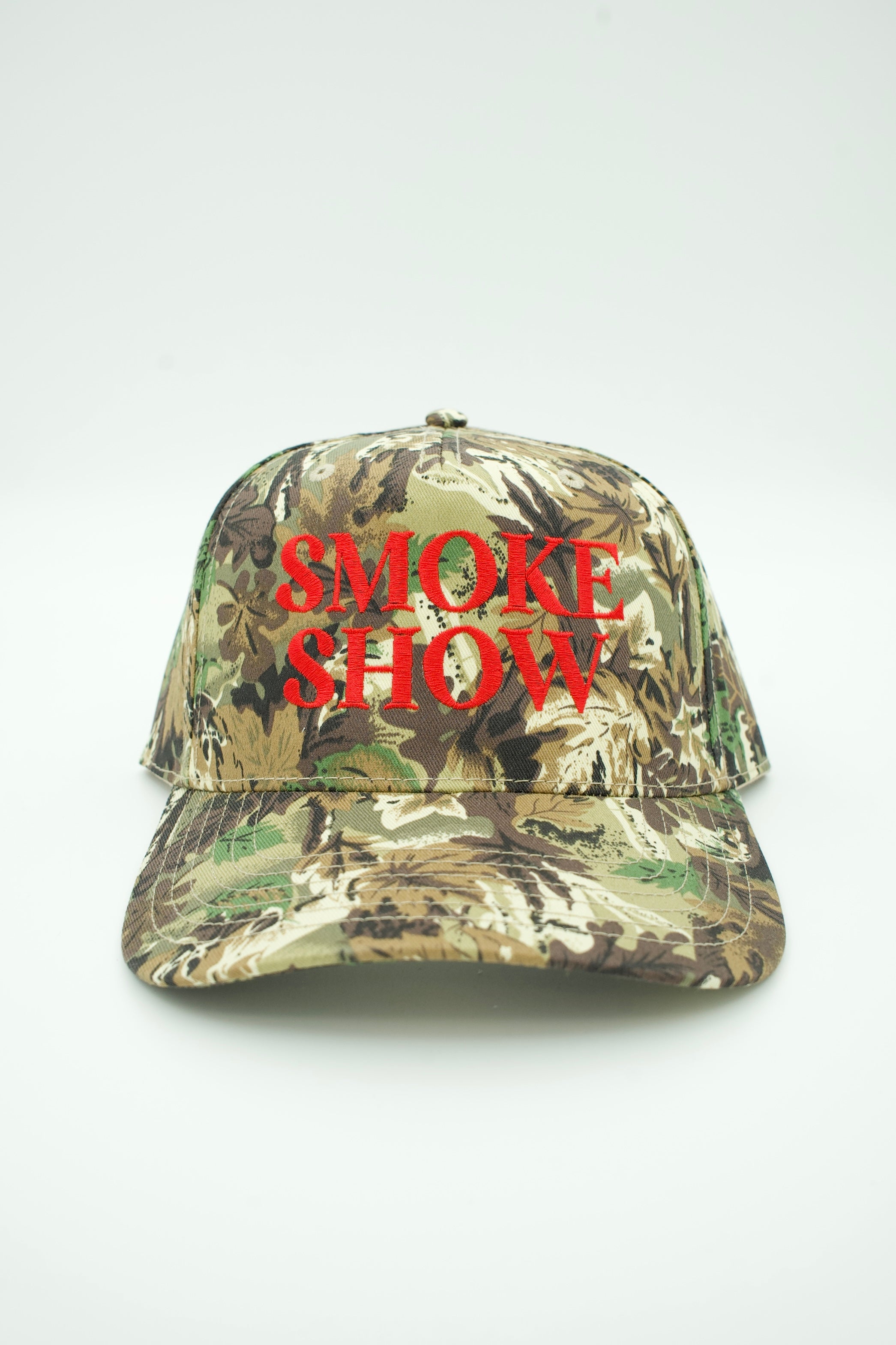 A camouflage baseball cap with the words 'SMOKE SHOW' embroidered in bold red letters on the front, set against a plain light gray background.