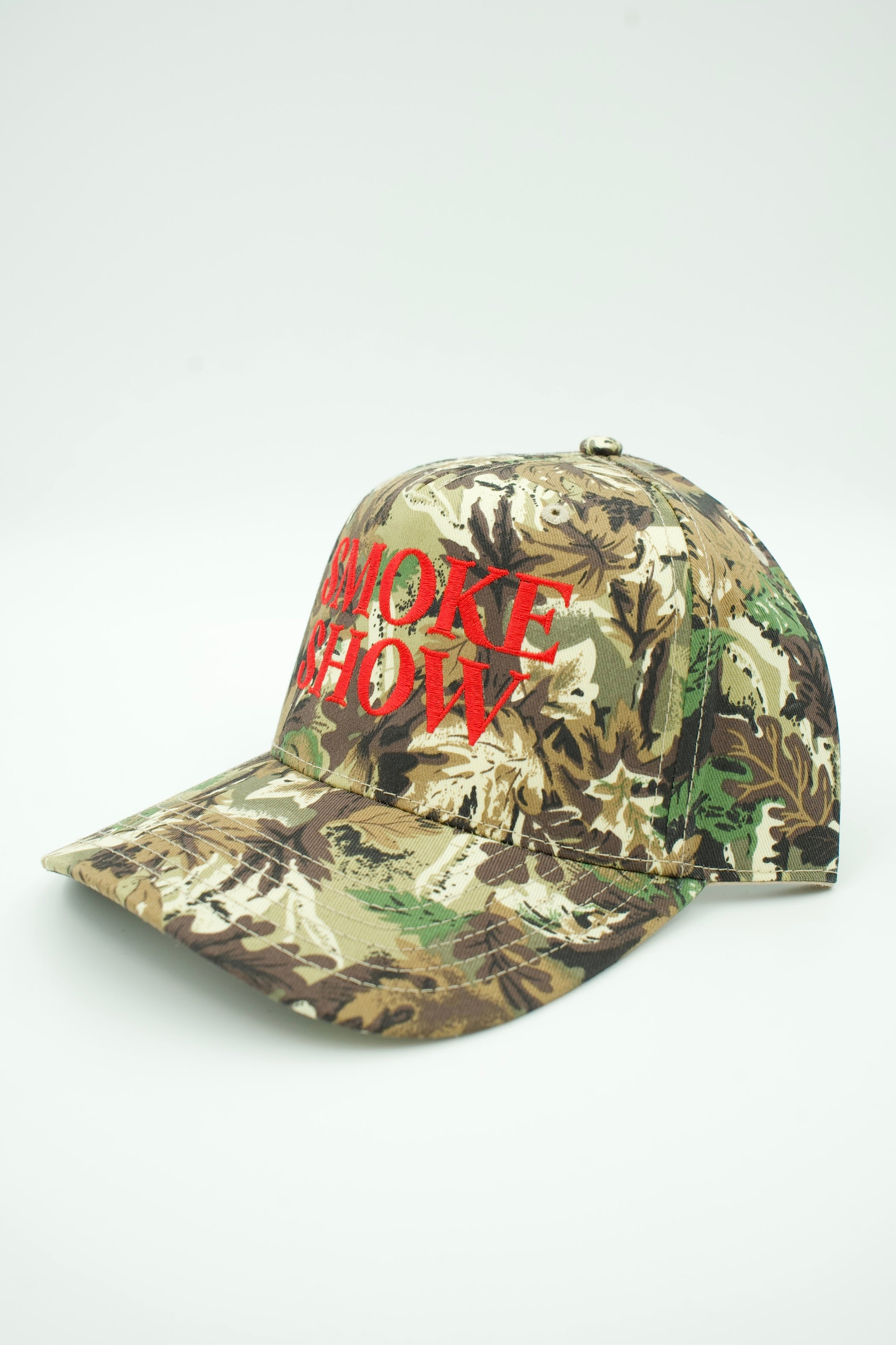 A baseball cap in a woodland camouflage pattern with red embroidered text reading 'SMOKE SHOW' on the front, photographed on a plain light background.