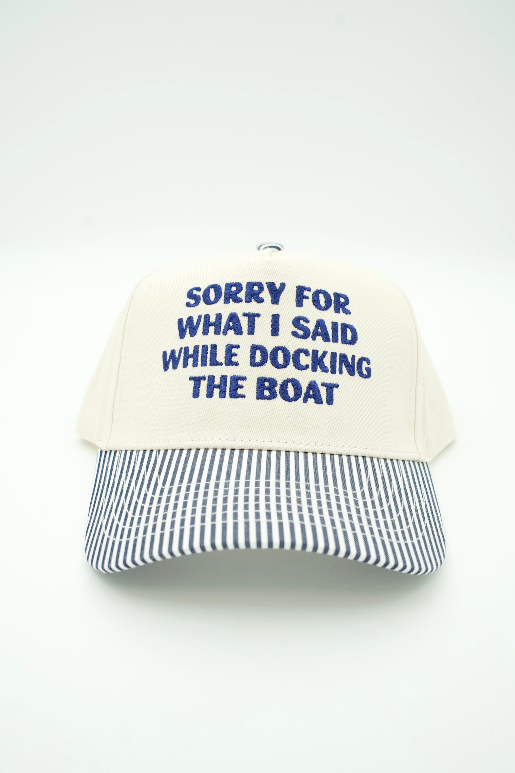 An off-white baseball cap with navy blue embroidered text reading 'SORRY FOR WHAT I SAID WHILE DOCKING THE BOAT' on the front, and a navy and white striped brim, shown against a plain white background.