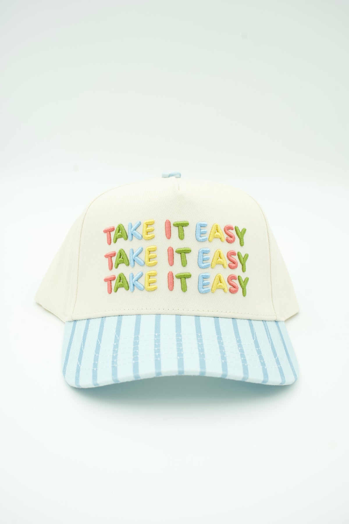 A baseball cap with an off-white crown, multicolored embroidered text reading 'TAKE IT EASY' repeated three times, and a light blue and white vertically striped brim, shown on a plain white background.