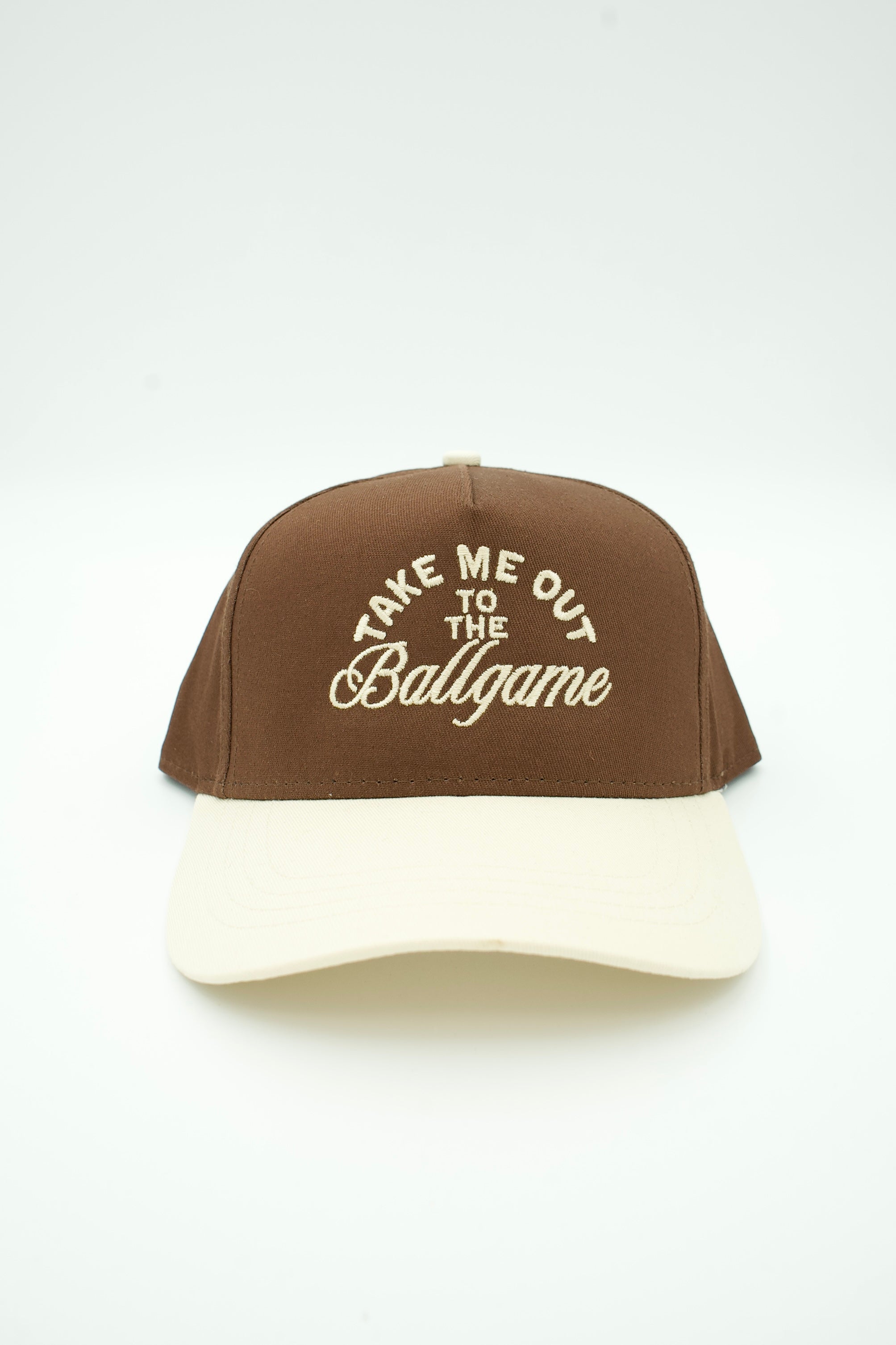 A brown baseball cap with a cream-colored visor and cream embroidered text on the front that reads 'TAKE ME OUT TO THE Ballgame'.