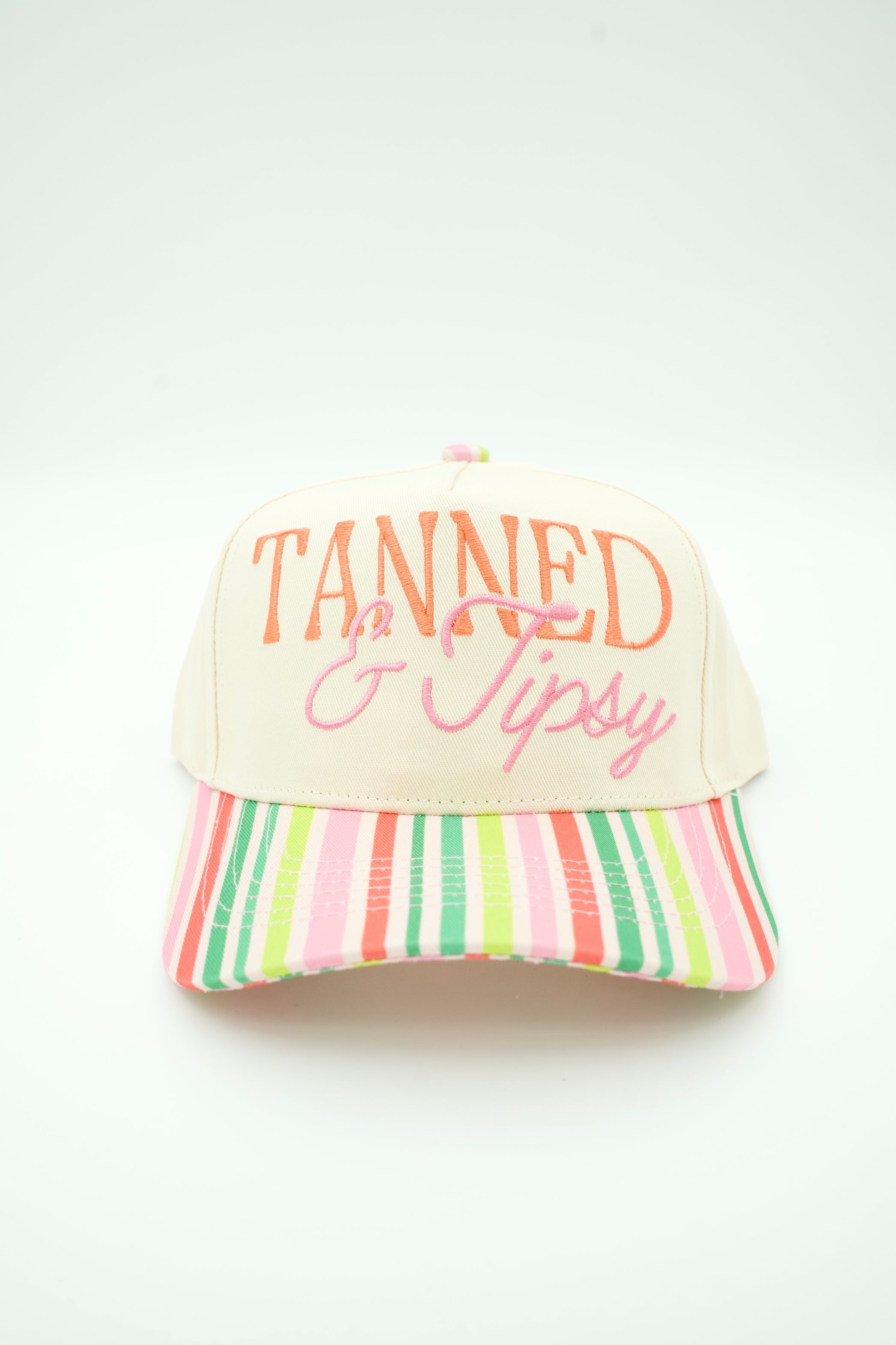 A baseball cap with a cream-colored front panel and brim, featuring the text 'TANNED & Tipsy' embroidered on the front in orange and pink. The brim is decorated with vertical pastel stripes in pink, green, orange, and white.